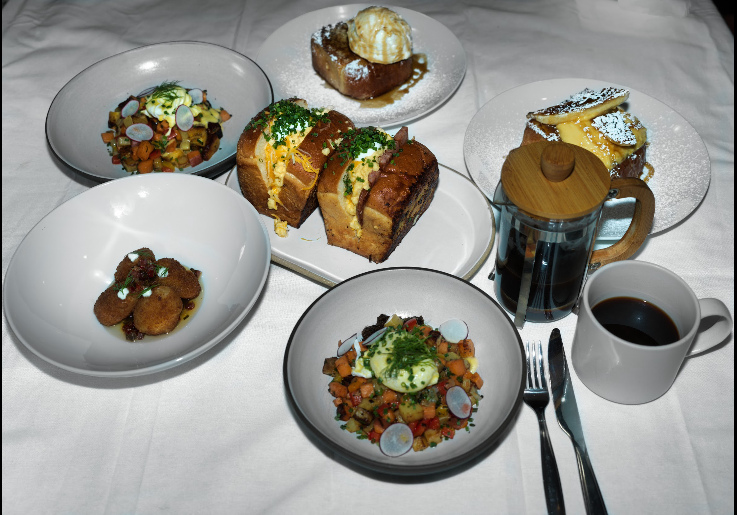 A table with various dishes including two plates of eggs, one plate with a vegetable hash, one plate with fried food, two dessert dishes with pastries and ice cream, along with a cup of coffee and a glass of soda.