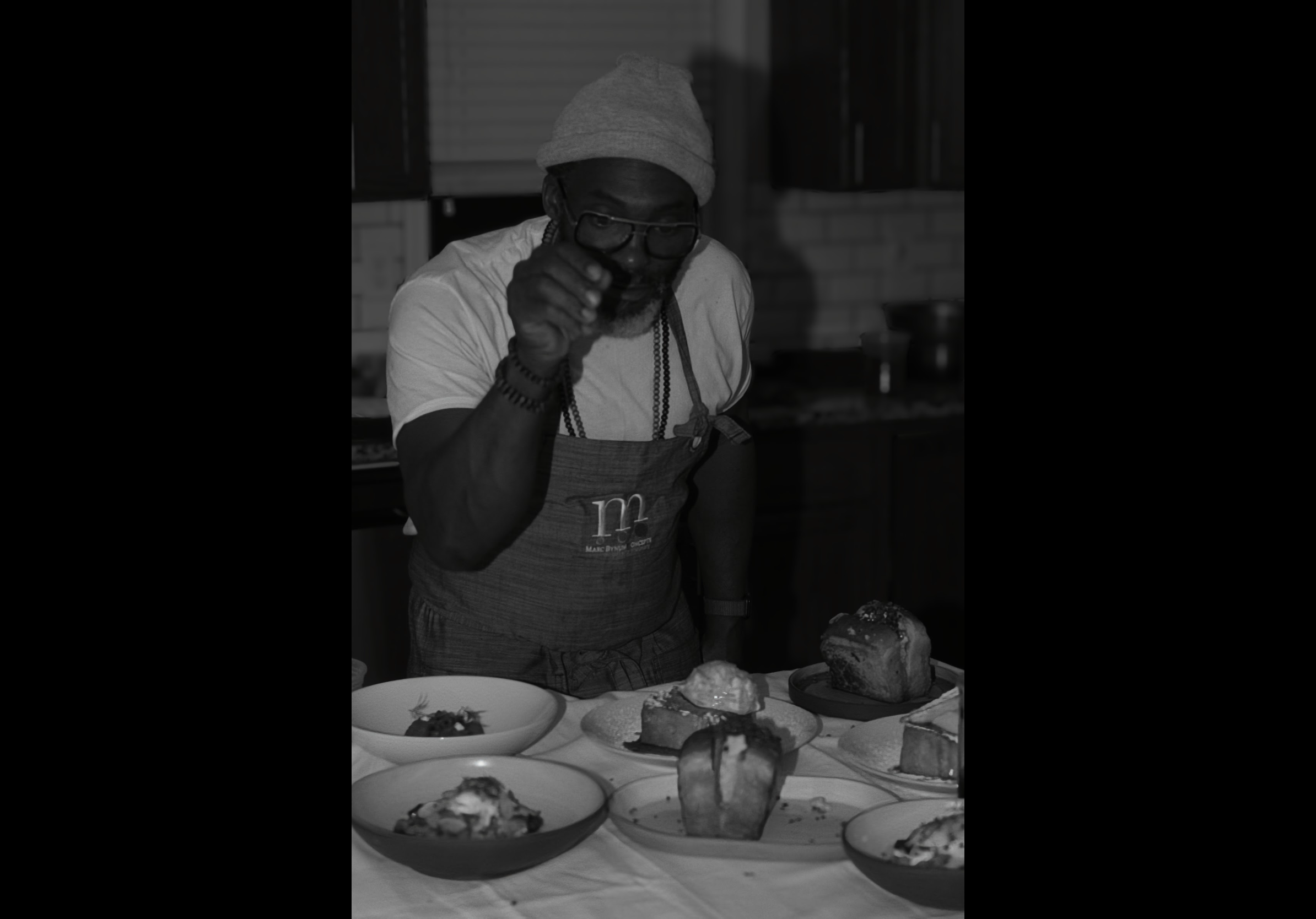 A man wearing glasses, a gray beanie, a white t-shirt, and jewelry is standing behind a table with various dishes, including bread, desserts, and small plates of food, in a kitchen or dining area.