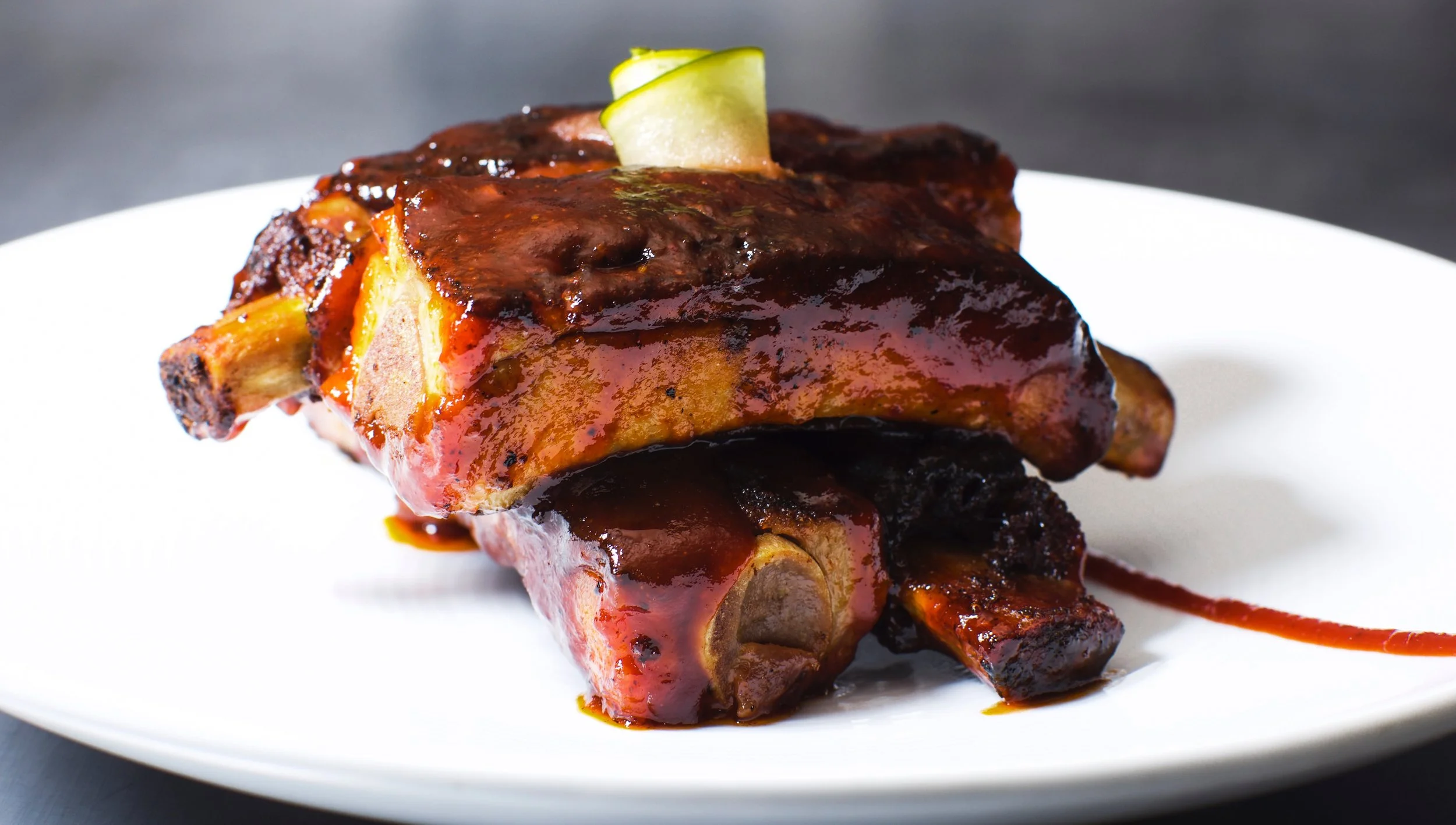 A close-up image of a serving of barbecued meat on a white plate, topped with barbecue sauce and garnished with a small piece of green onion.