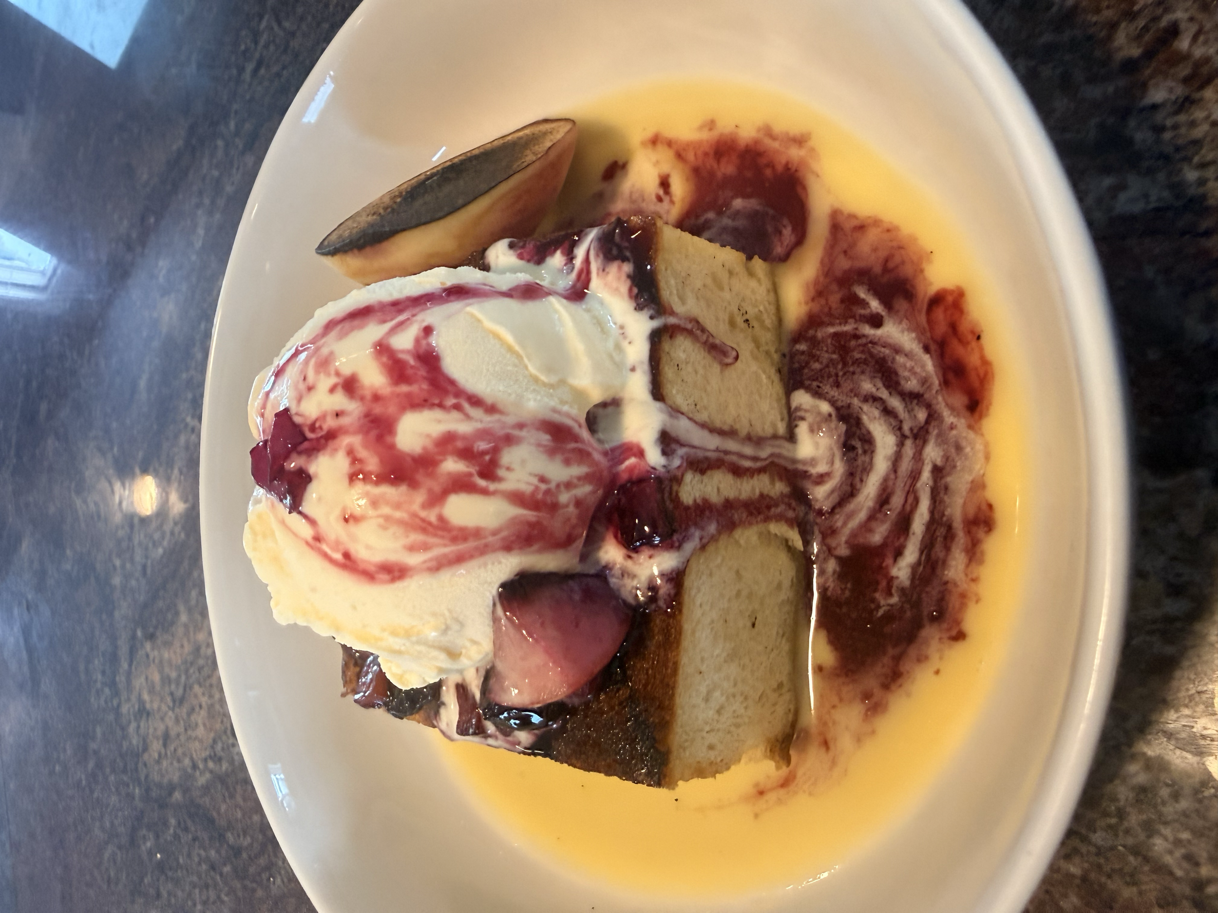 A dessert with a slice of cake topped with vanilla ice cream, berry compote, and a slice of banana, served in a white bowl with a sauce at the bottom.