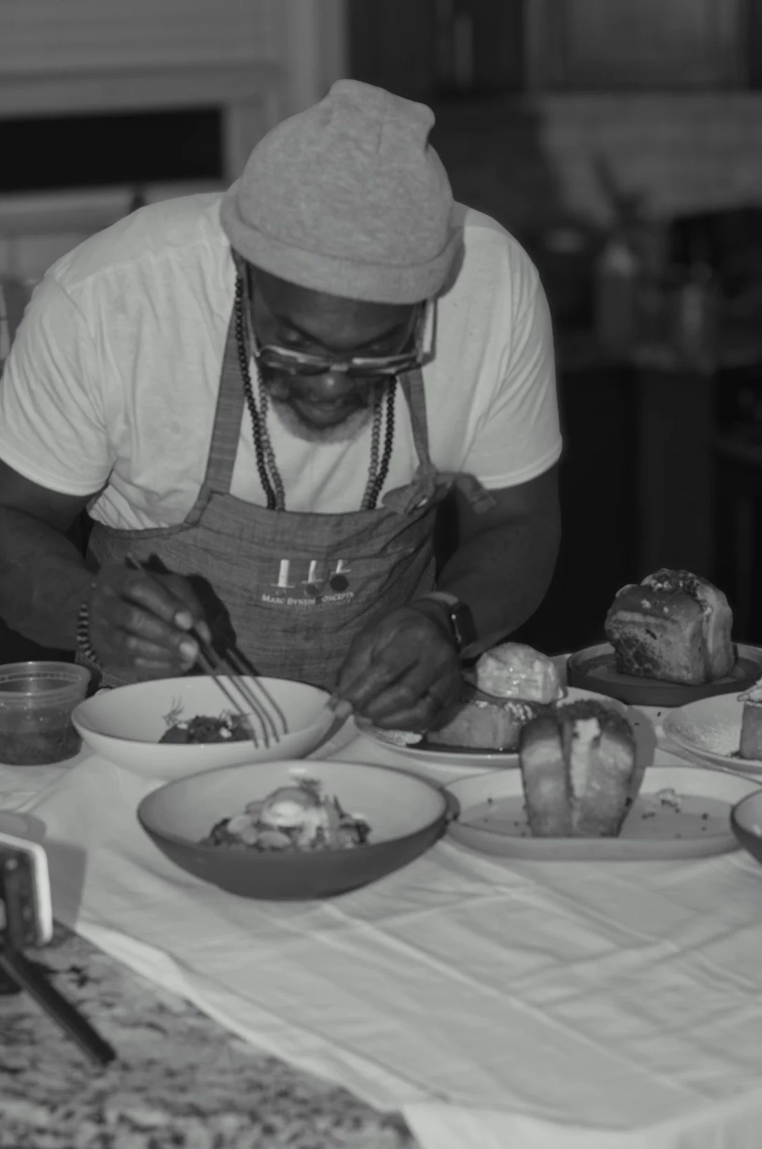 A man wearing a beanie, glasses, and an apron preparing food at a table with various dishes and bread in a kitchen or dining area.