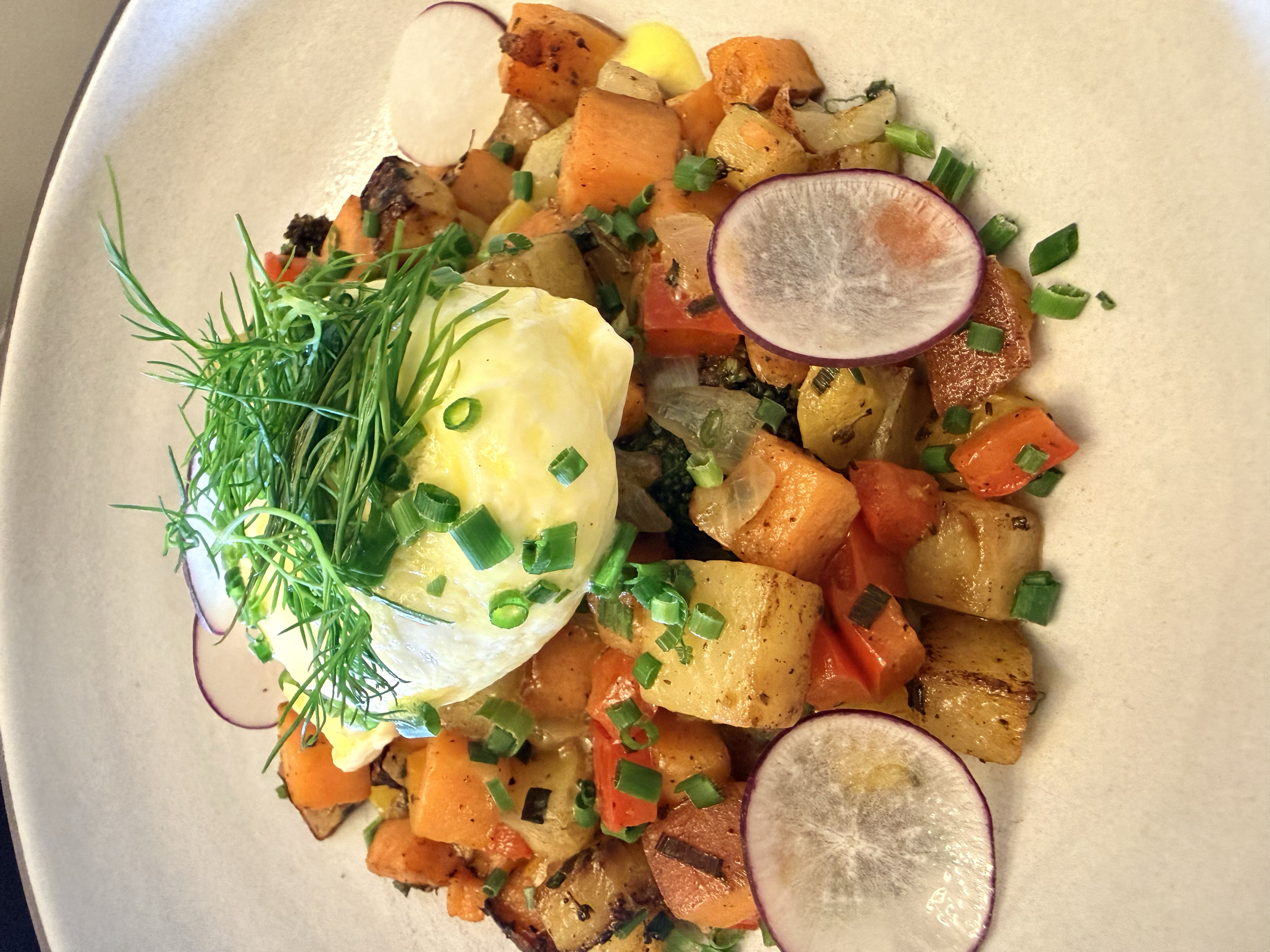 A plate of roasted sweet potatoes, carrots, and potatoes topped with a poached egg, chopped green onions, radish slices, and garnished with fresh herbs.
