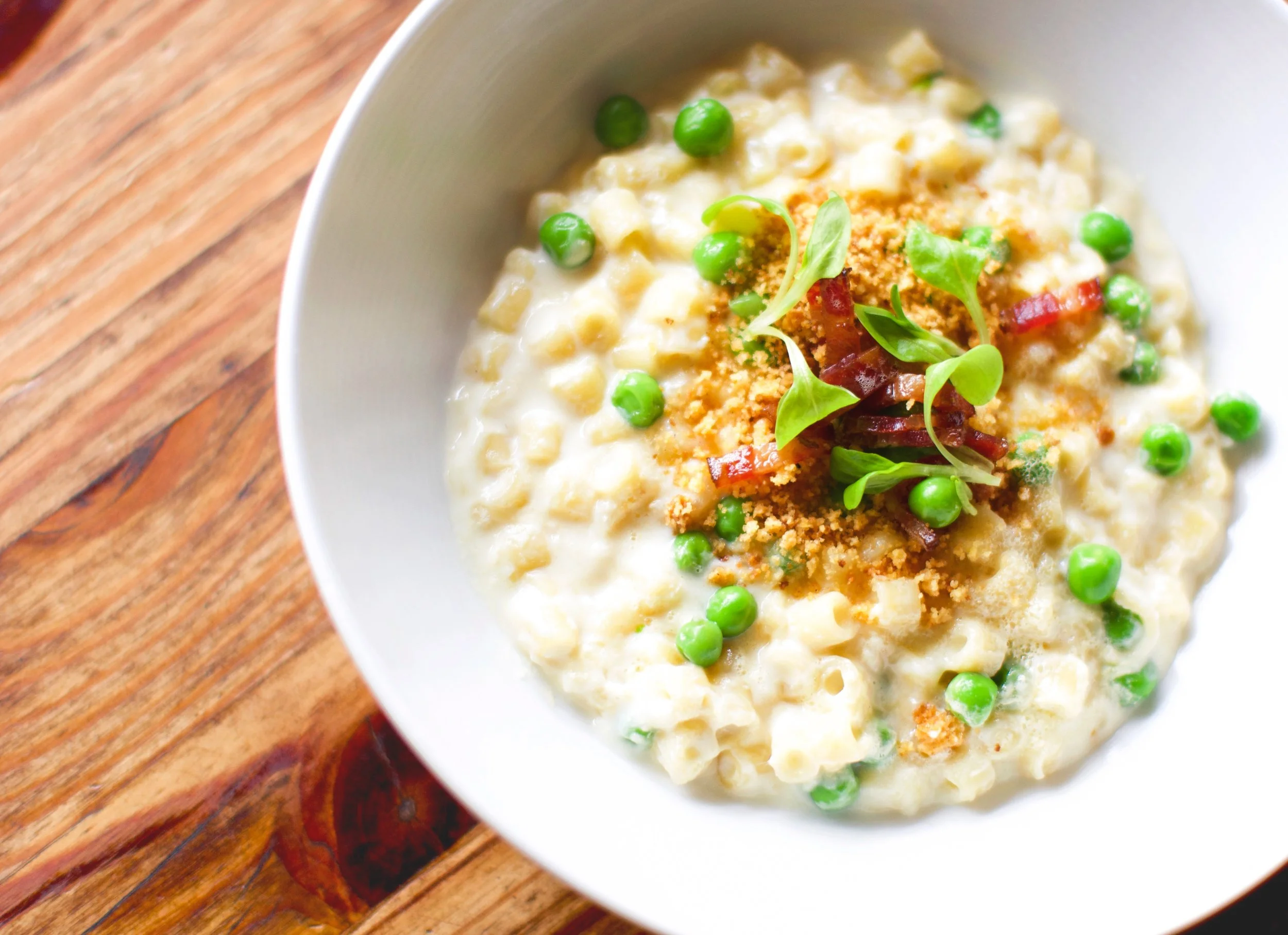 Close-up of a bowl of creamy macaroni and cheese topped with green peas, crispy bacon bits, and chopped herbs on a wooden table.