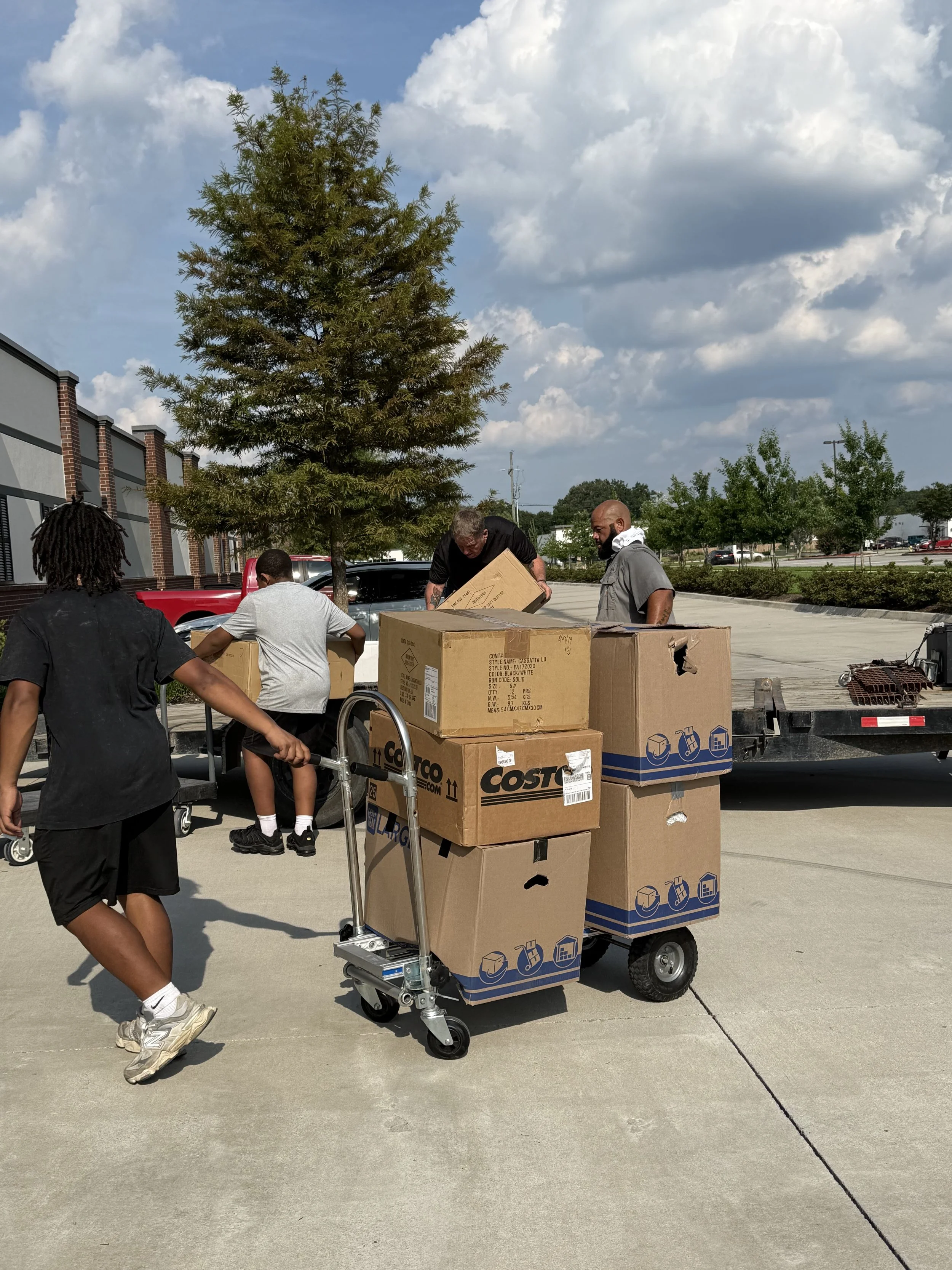 People unloading and organizing boxes in a parking lot with a large tree and cloudy sky in the background.