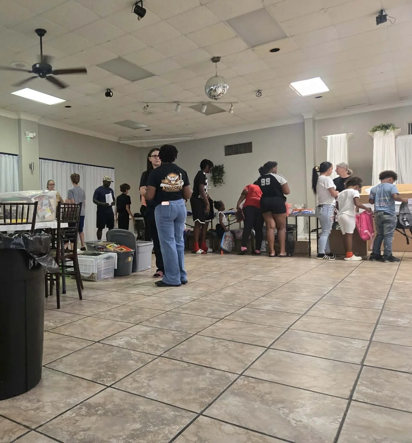 People standing in line at a table in an indoor event space with a tiled floor, ceiling fans, and a disco ball.