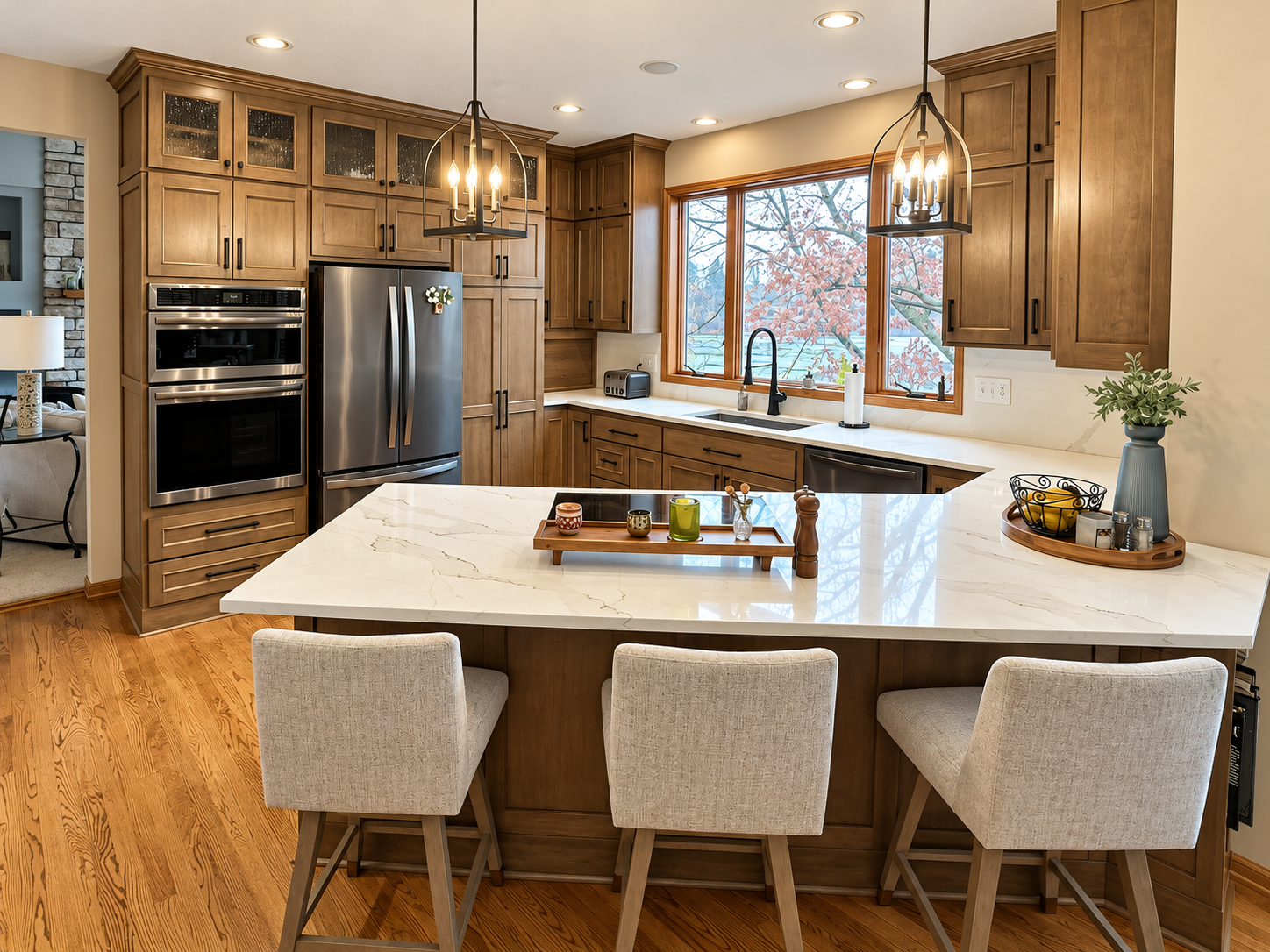 Modern kitchen with white marble island, wooden cabinets, stainless steel appliances, and large window showing trees outside. Four beige chairs are at the island, with decorative candles and plants on the countertop and tray.