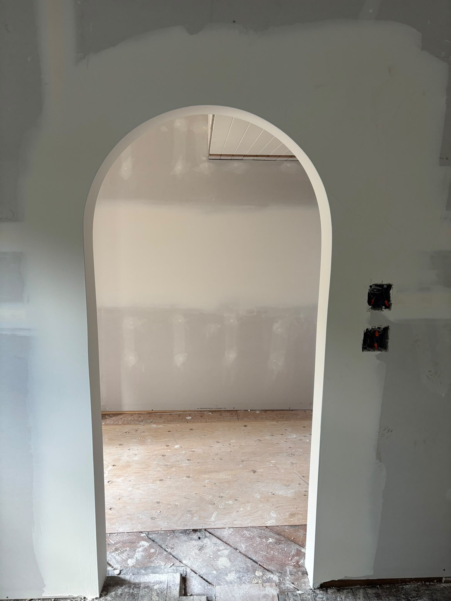 A partially finished room under construction with an archway. The walls are unpainted drywall, and electrical outlets are exposed with wiring visible.
