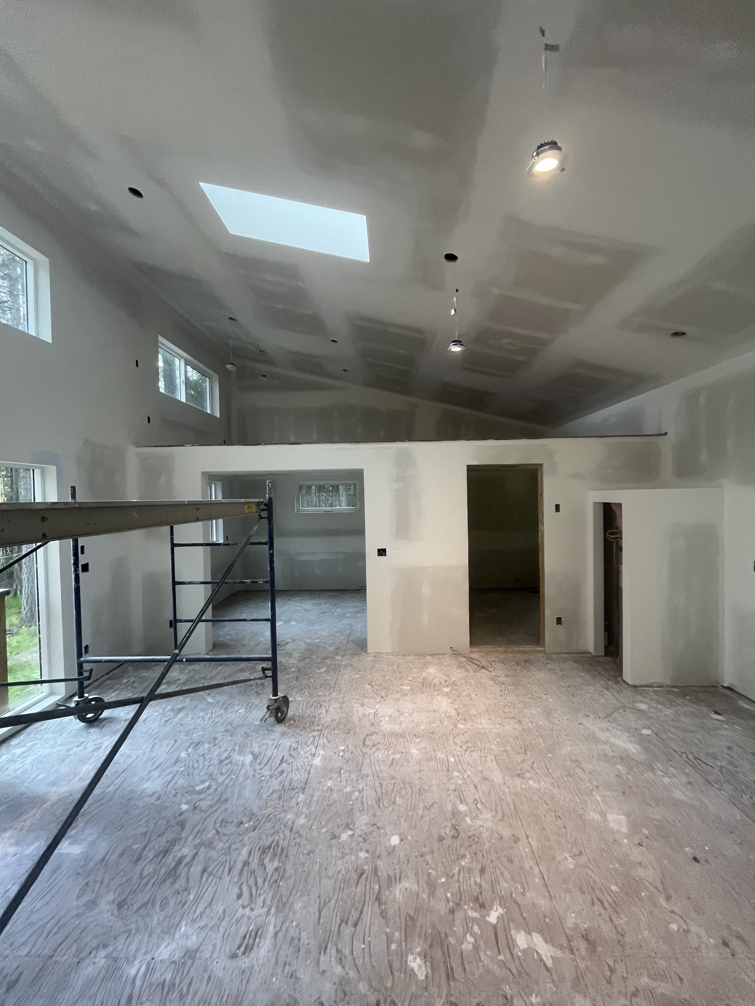 Interior of a house under construction with drywall and ceiling work being done, scaffolding visible, large windows, and unfinished flooring.