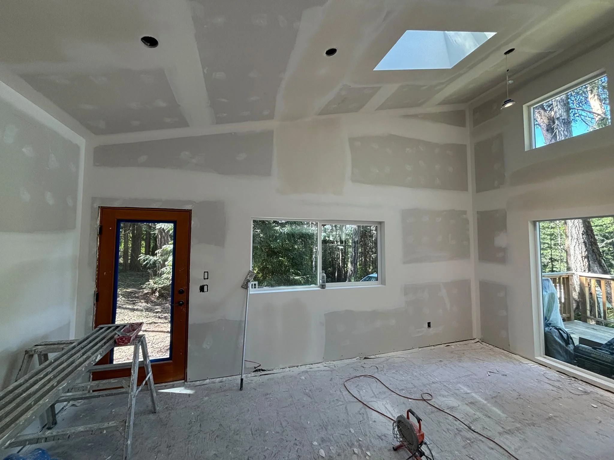 Interior of a house under construction with drywall installed on walls and ceiling, windows, a door, and construction tools and materials on the floor.
