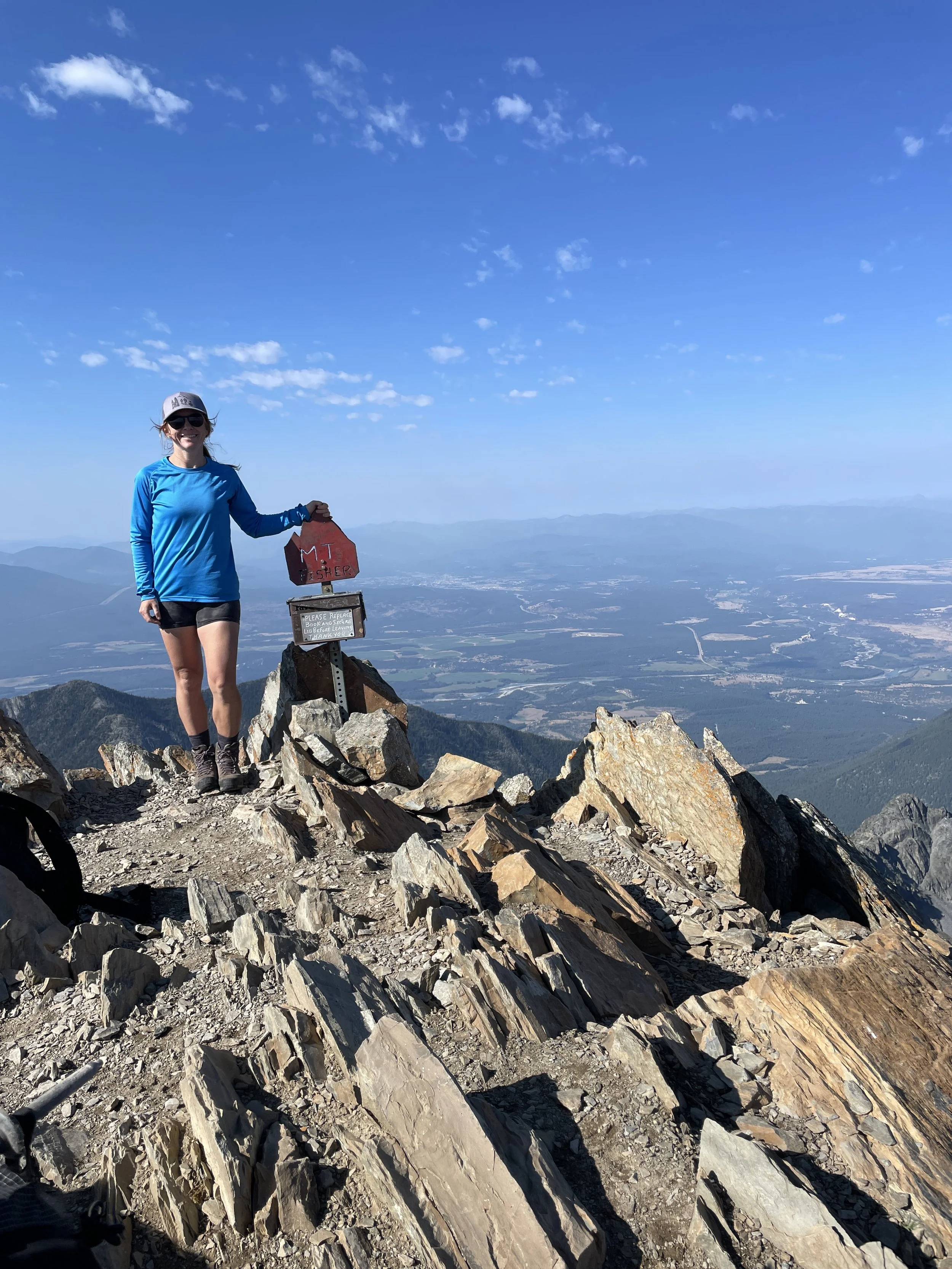 A woman dressed in outdoor hiking gear standing on a rocky mountain summit with a sign that says 'Mt Fisher'. She is smiling, wearing sunglasses and a cap, with a vast landscape of valleys and mountains in the background under a clear blue sky.