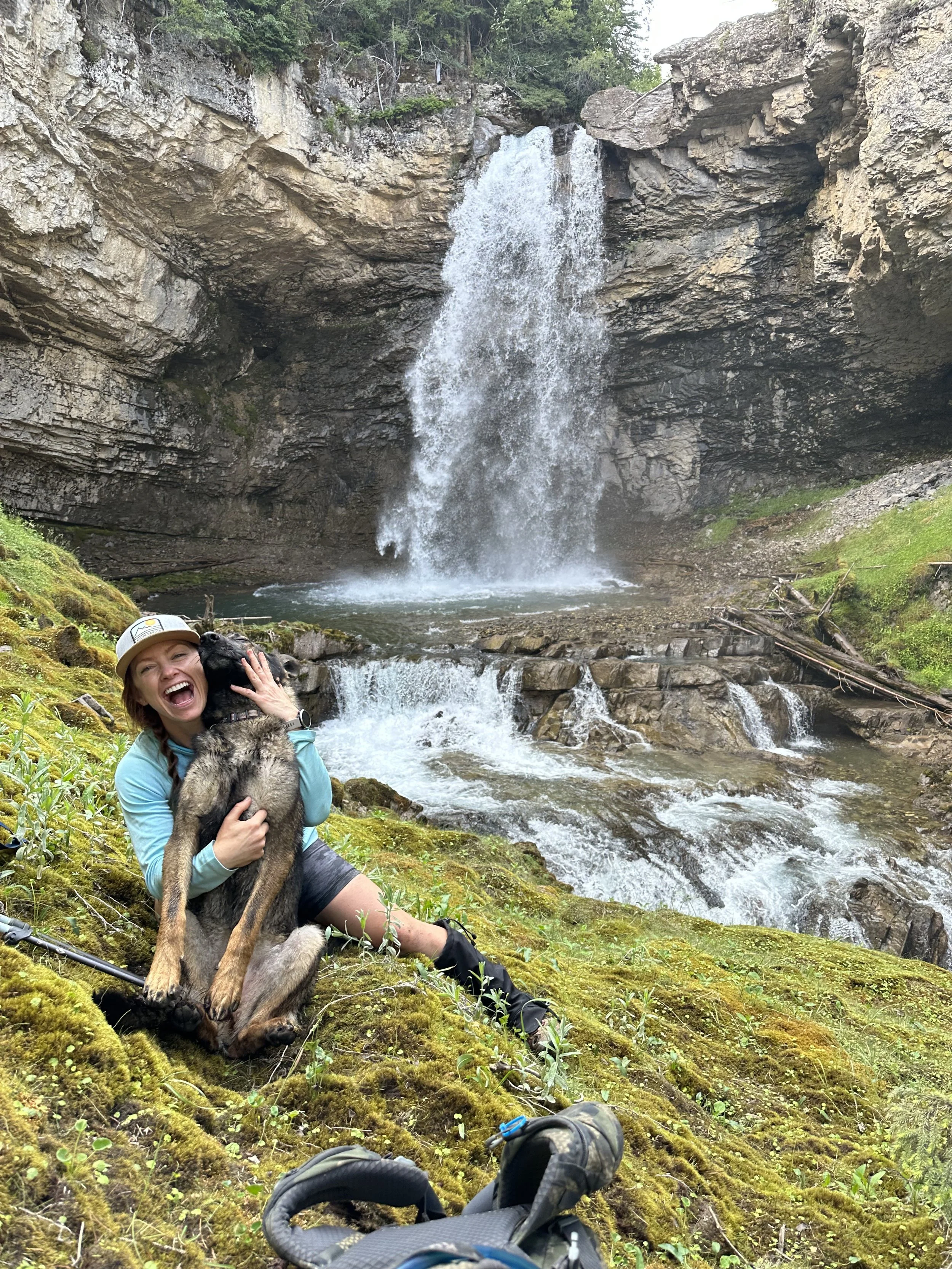A woman sitting on mossy ground by a river with a large waterfall in the background, holding a black and tan dog, smiling and laughing, with a pair of hiking shoes and a blue backpack in the foreground.
