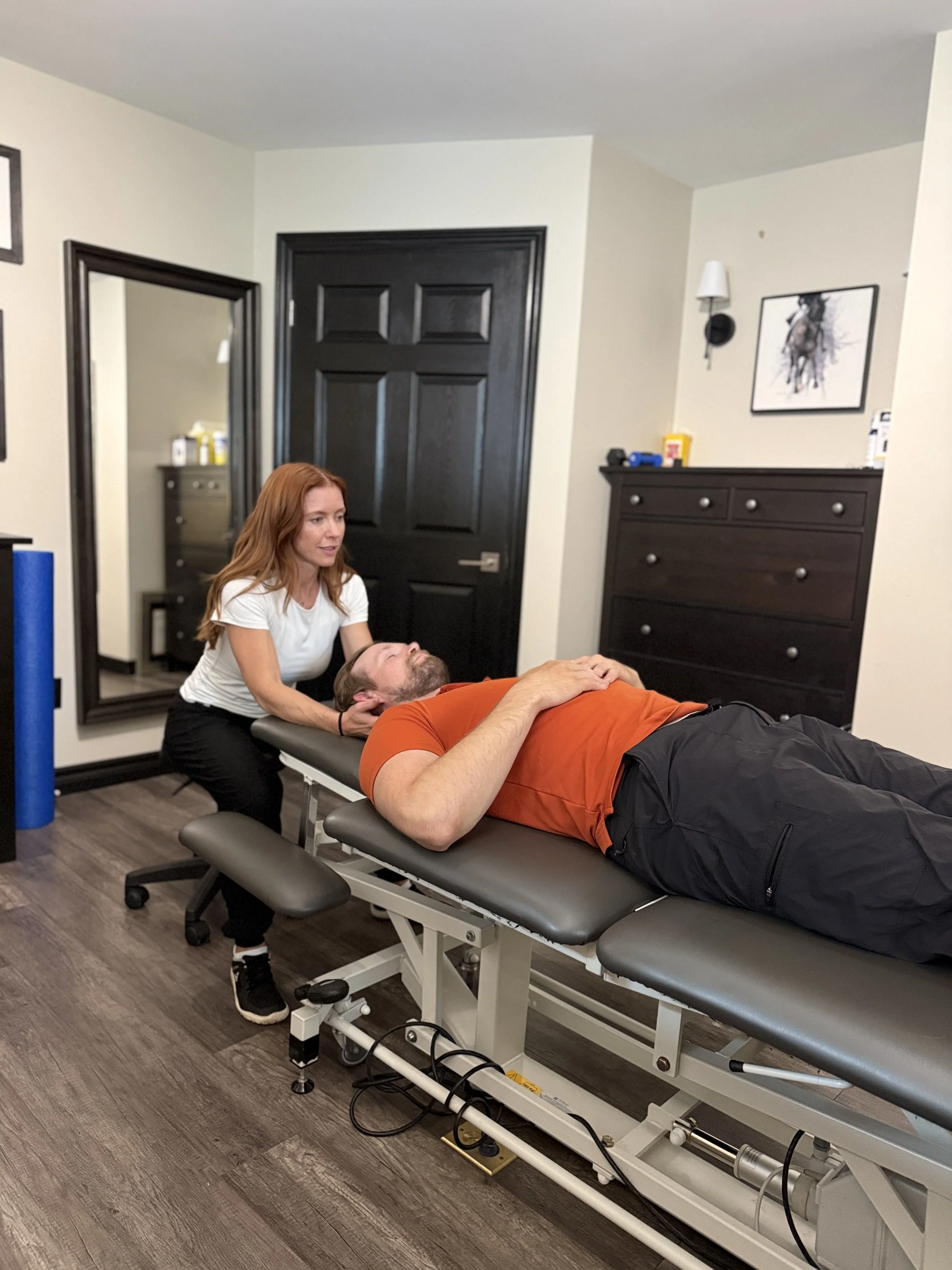 A woman with red hair and a white shirt performs a chiropractic adjustment on a man lying face up on a chiropractic table at a clinic or medical office.