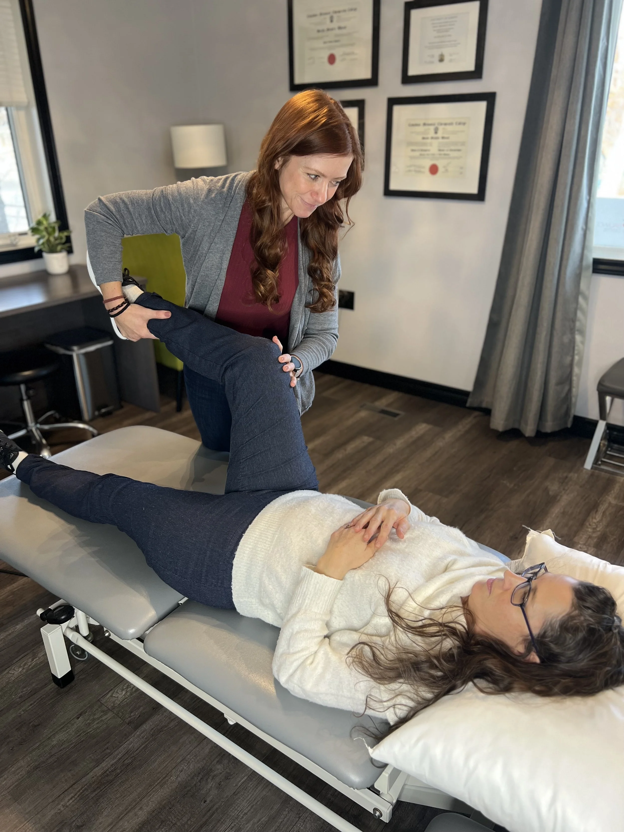 A woman with glasses lying on a medical examination table with her eyes closed, receiving manual therapy from a chiropractor who is holding her leg. The room has framed certificates on the wall and windows with curtains.