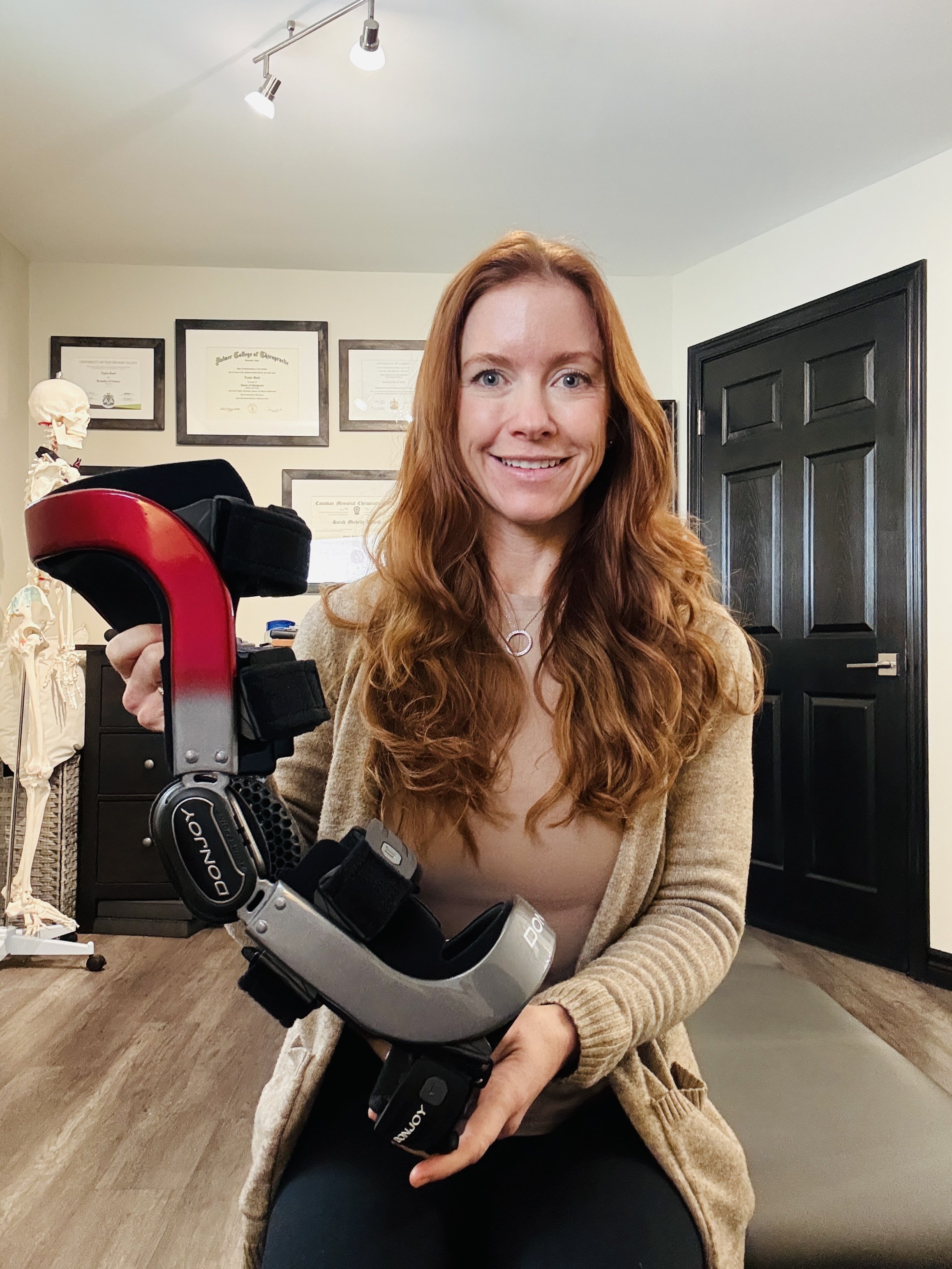 A woman with long curly red hair smiling and holding a custom knee brace in a room with framed certificates on the wall, a skeleton model, and a black door.