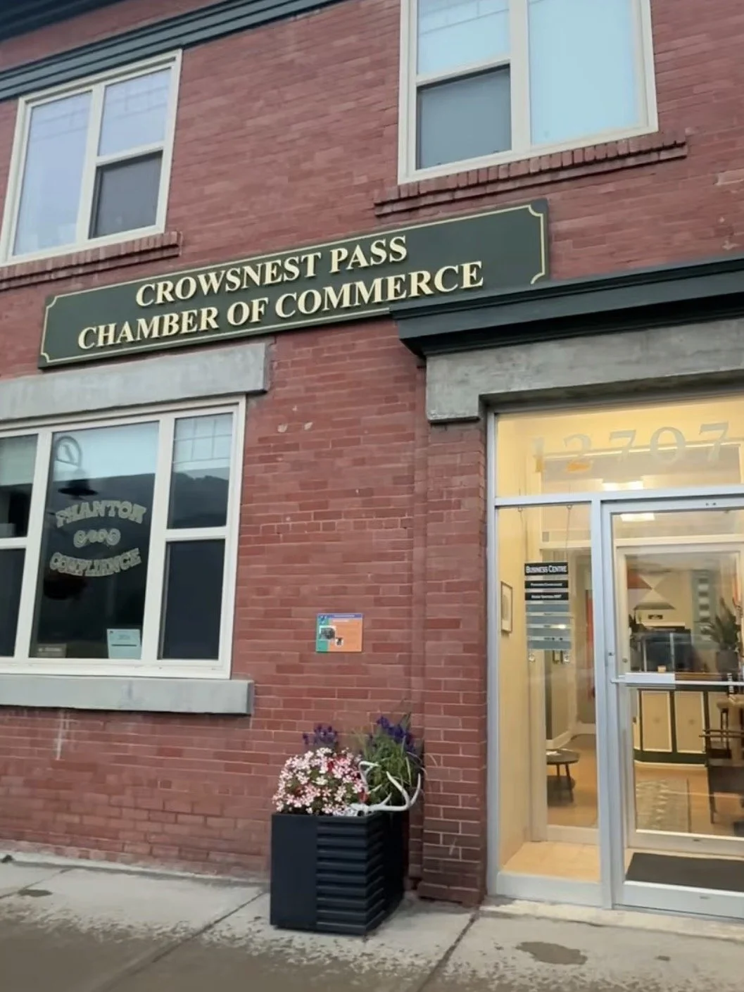 Red brick building with a green sign that reads 'Crowsnest Pass Chamber of Commerce,' and a glass door entrance with the address 12207. There is a black planter with pink and purple flowers outside.