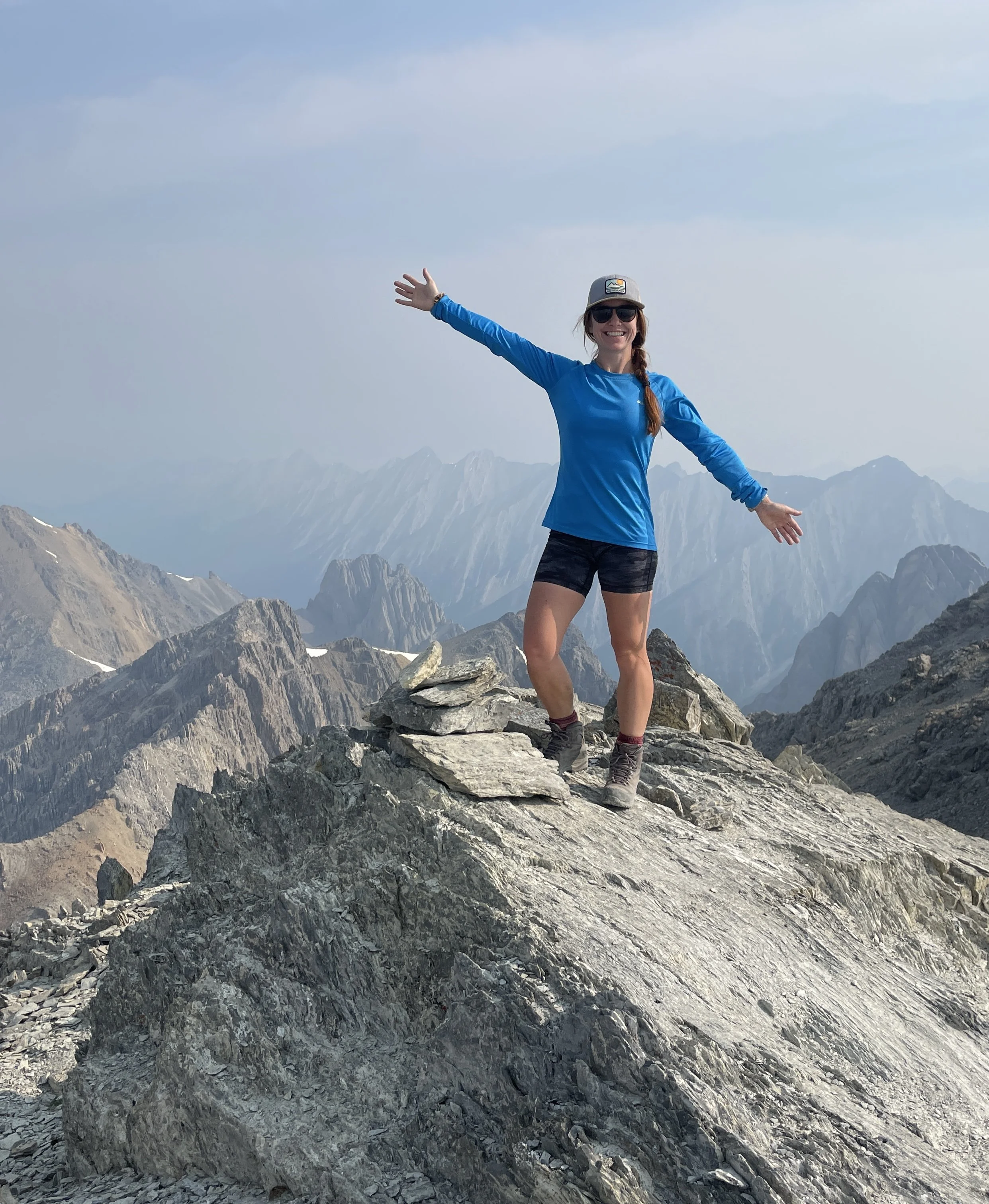 A woman standing on rocky mountain terrain with jagged peaks in the background. She is wearing a blue long-sleeve shirt, black shorts, hiking boots, a cap, and sunglasses, with arms outstretched and smiling.
