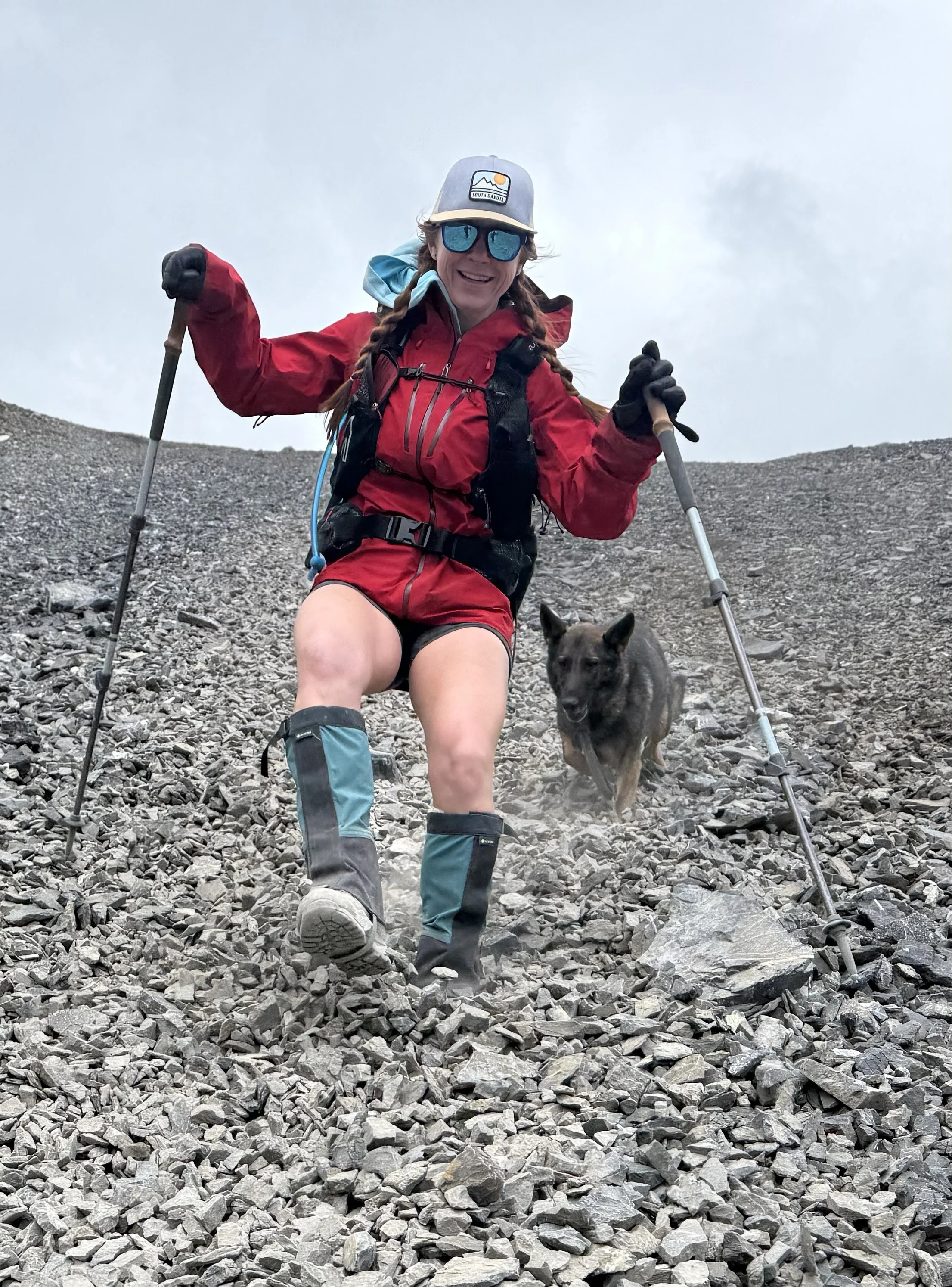 A woman hiking on a rocky terrain with a dog, wearing red outdoor gear, sunglasses, and a cap, with trekking poles, smiling at the camera.