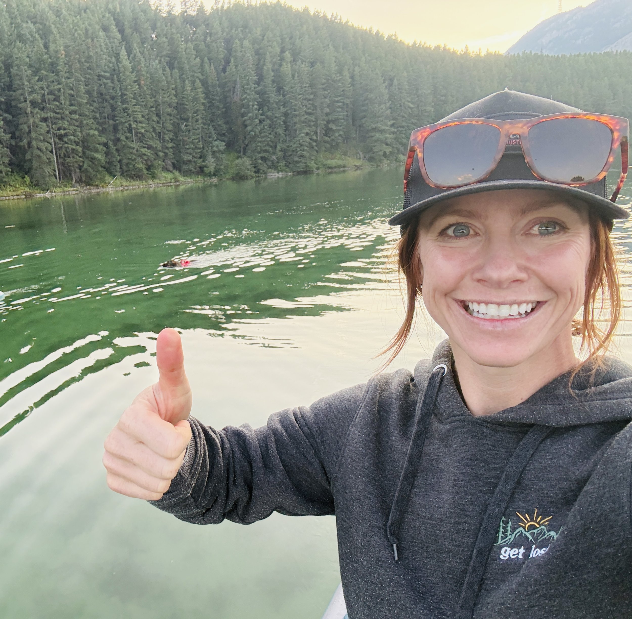 A person in a black hoodie with sunglasses resting on their head giving a thumbs up while on a paddle board in a lake surrounded by green forested mountains at sunset.
