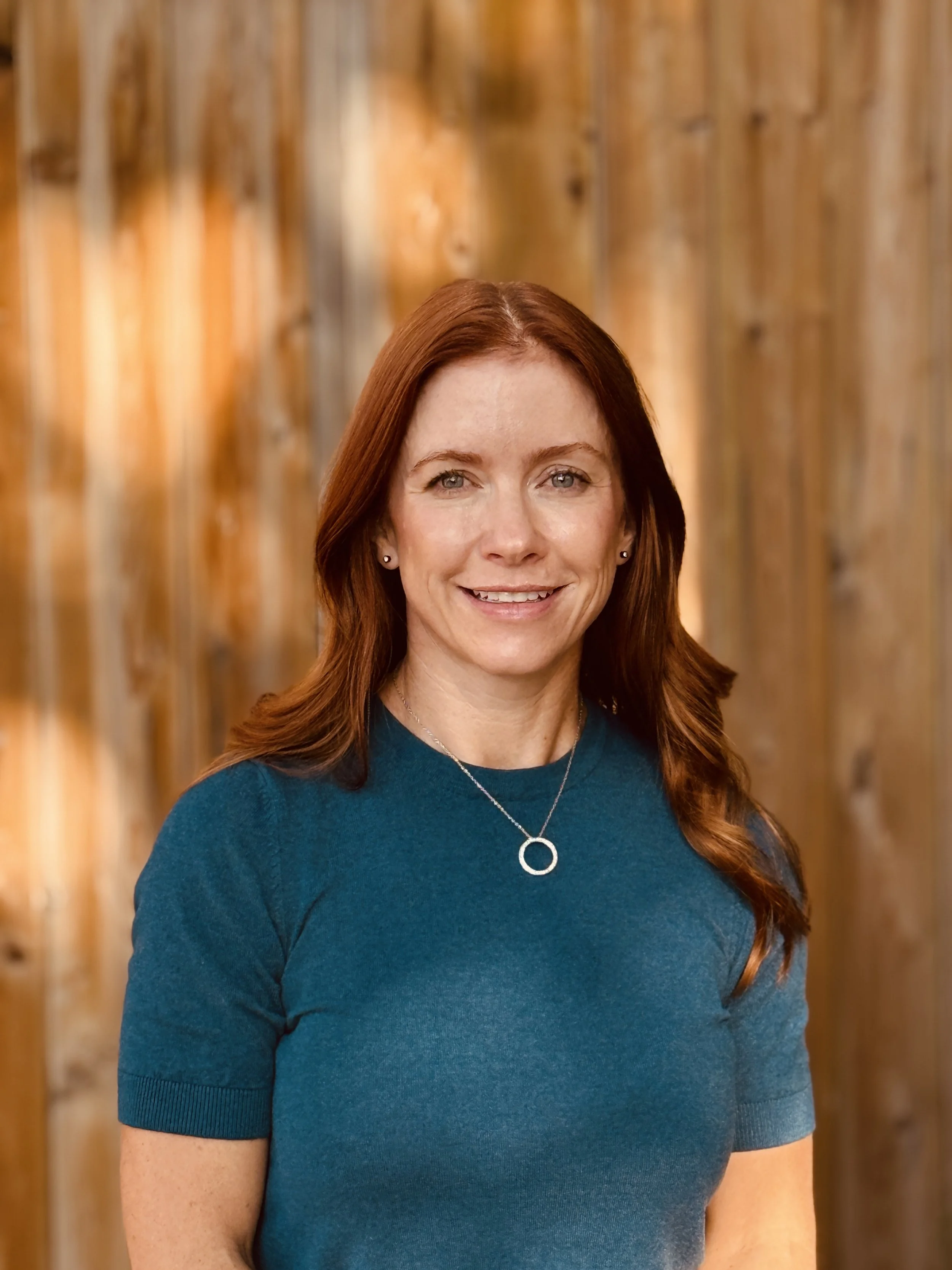 Portrait of a woman with red hair wearing a dark teal top and a silver pendant necklace, smiling, standing in front of a wooden background.