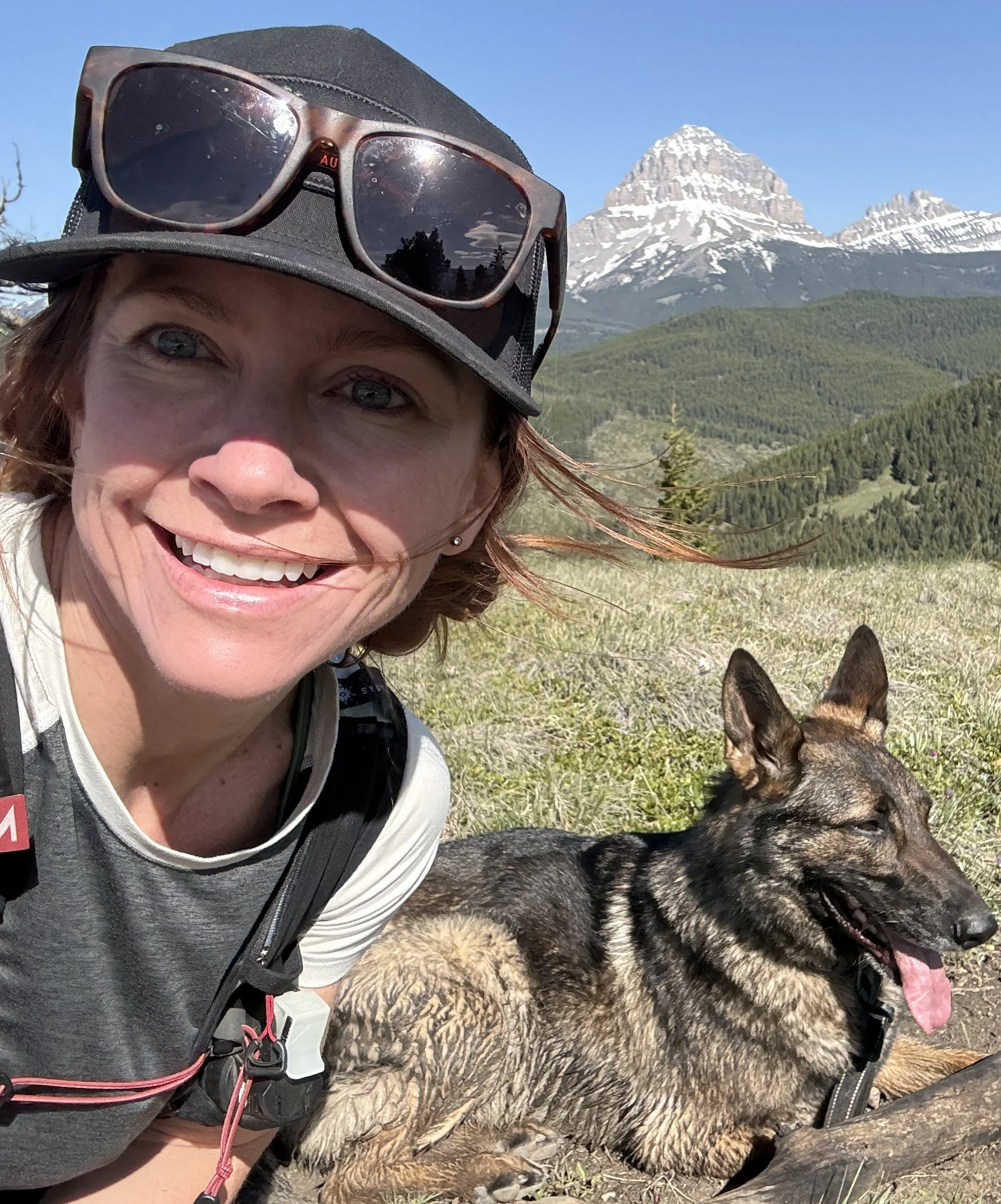 A smiling woman with sunglasses on her hat and a backpack poses with a large, fluffy German shepherd dog with a mountain range in the background, including snow-capped peaks and a clear blue sky.