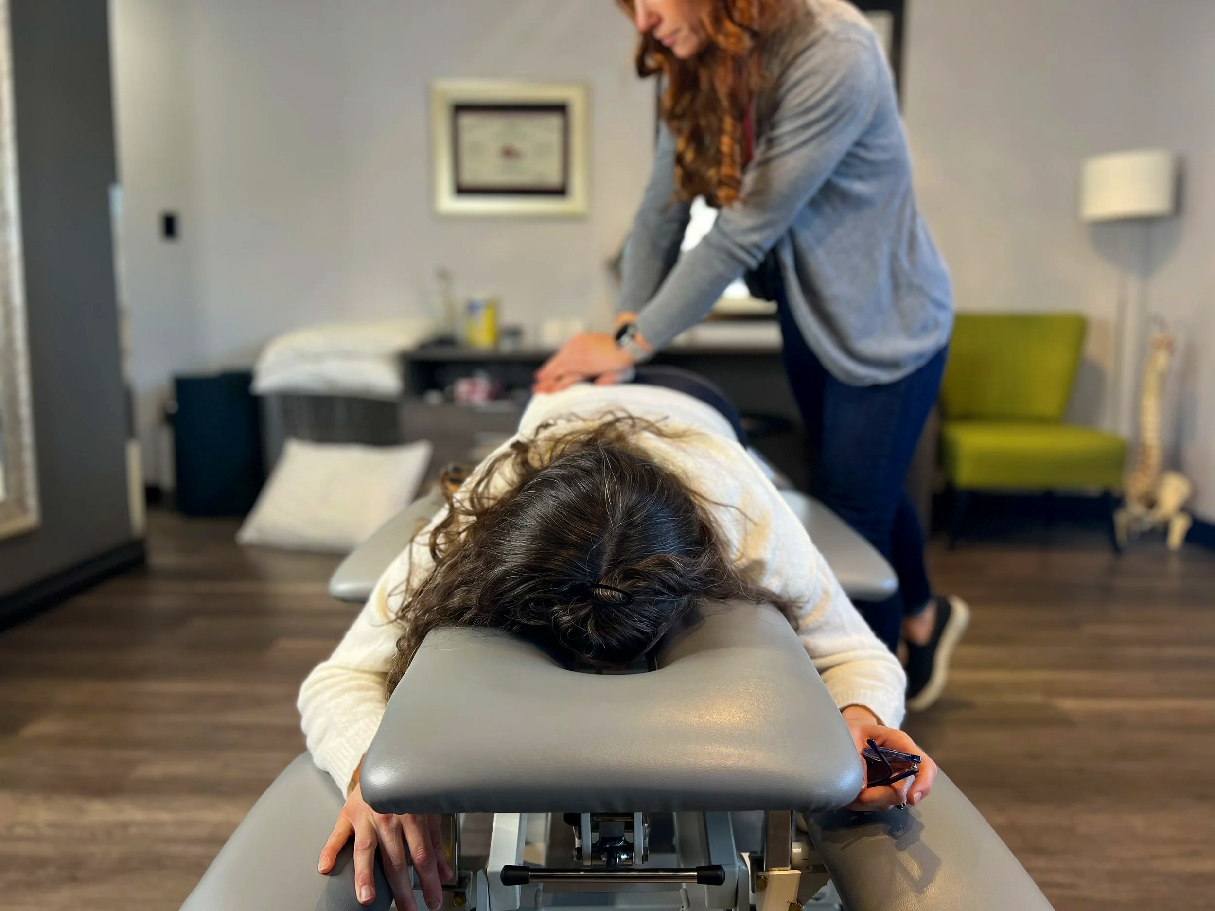 A woman lying face down on a chiropractic table with her eyes closed while the chiropractor applies pressure to her back with her hands in a chiropractic care session in a modern treatment room.
