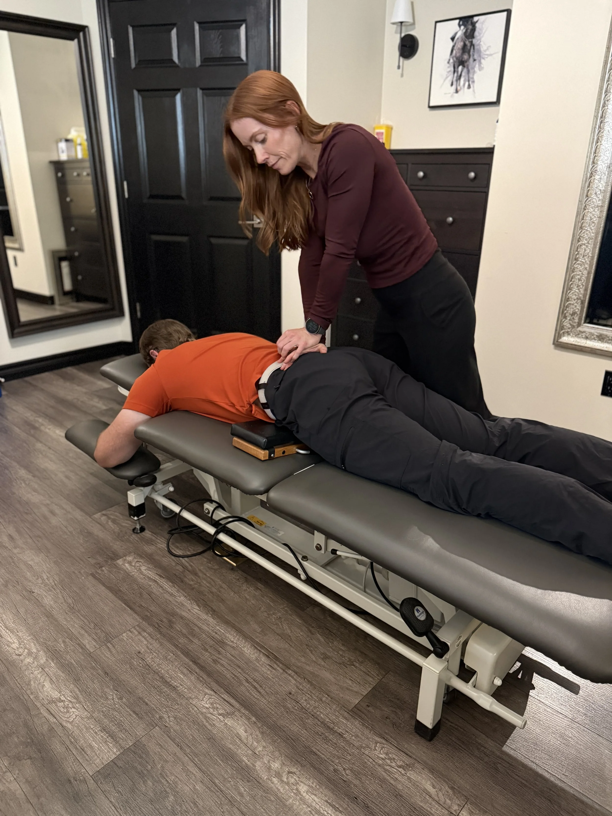 A woman performing an adjustment on a man lying face down on a chiropractic table in a room with wood flooring, a black door, and framed artwork on the wall.