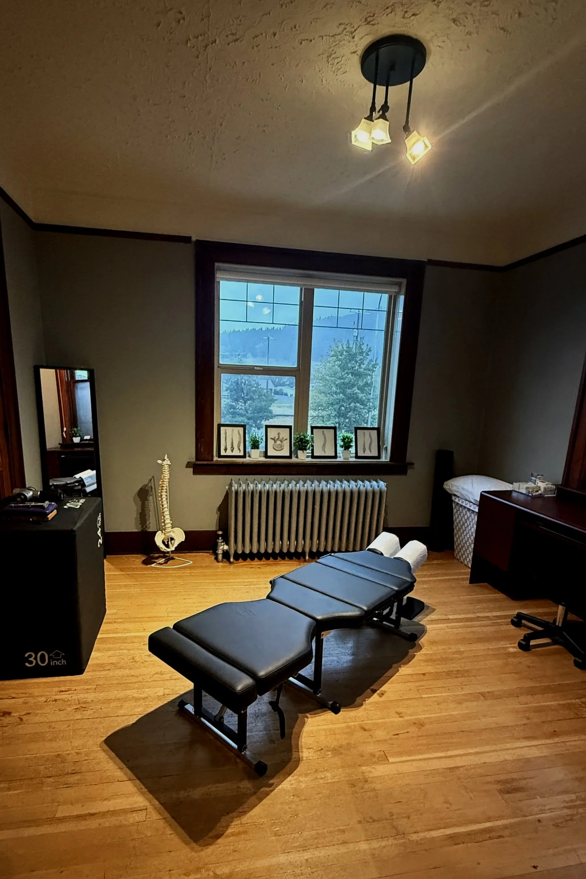 A room with wooden flooring, a large window with a mountain view, a massage table, a black speaker, a mirror, a small dresser, framed art on the windowsill, and a nearby music stand.