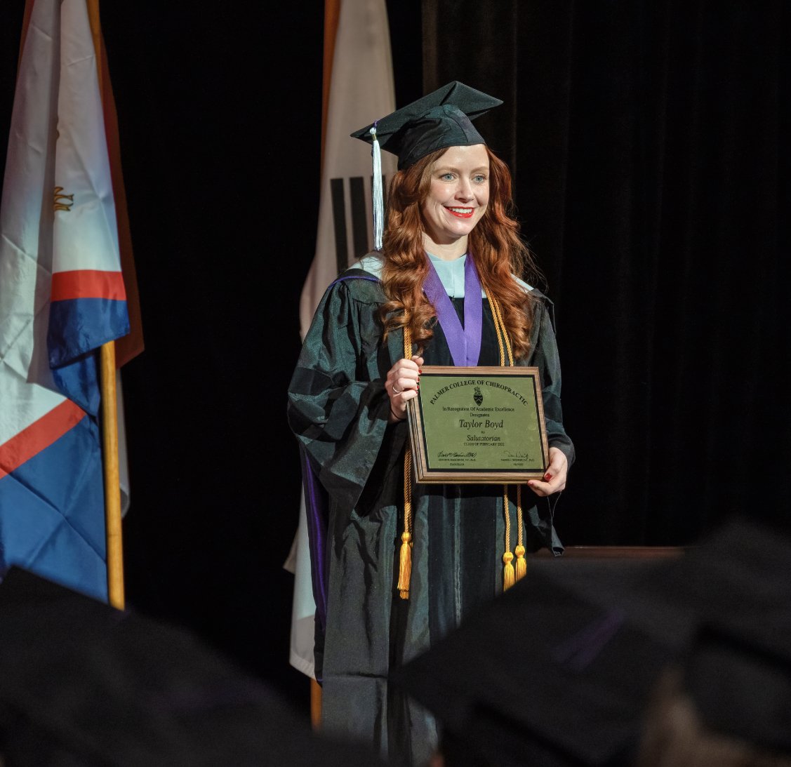 A woman with red hair in graduation cap and gown holding a diploma, smiling on stage, with flags in the background.