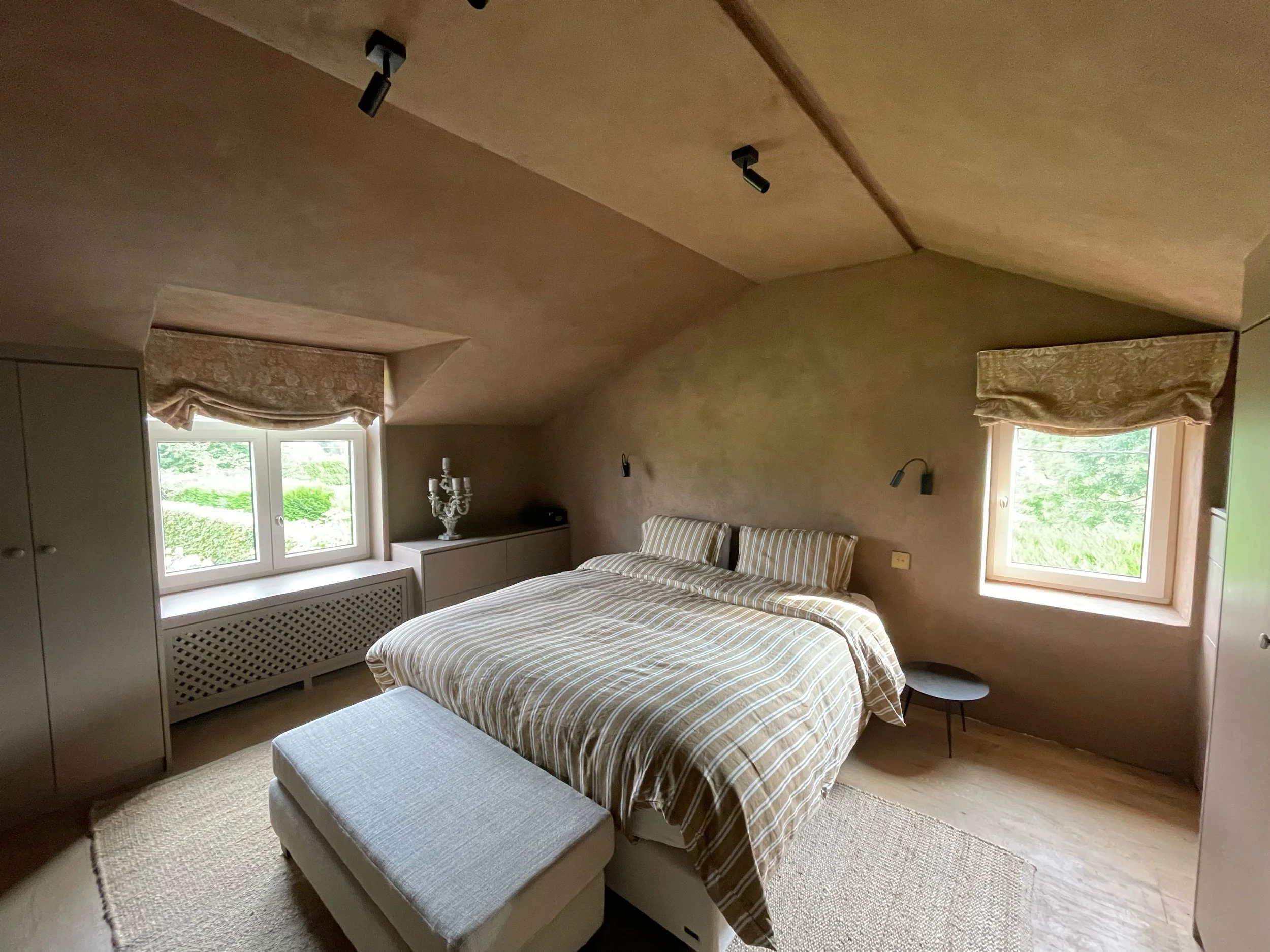 A cozy bedroom with a bed covered in beige and white striped bedding, positioned by two windows with beige curtains. There is a gray ottoman at the foot of the bed, a small black side table, and a white dresser with a decorative candelabra on top. The ceiling features a slightly curved design with modern black adjustable spotlights.