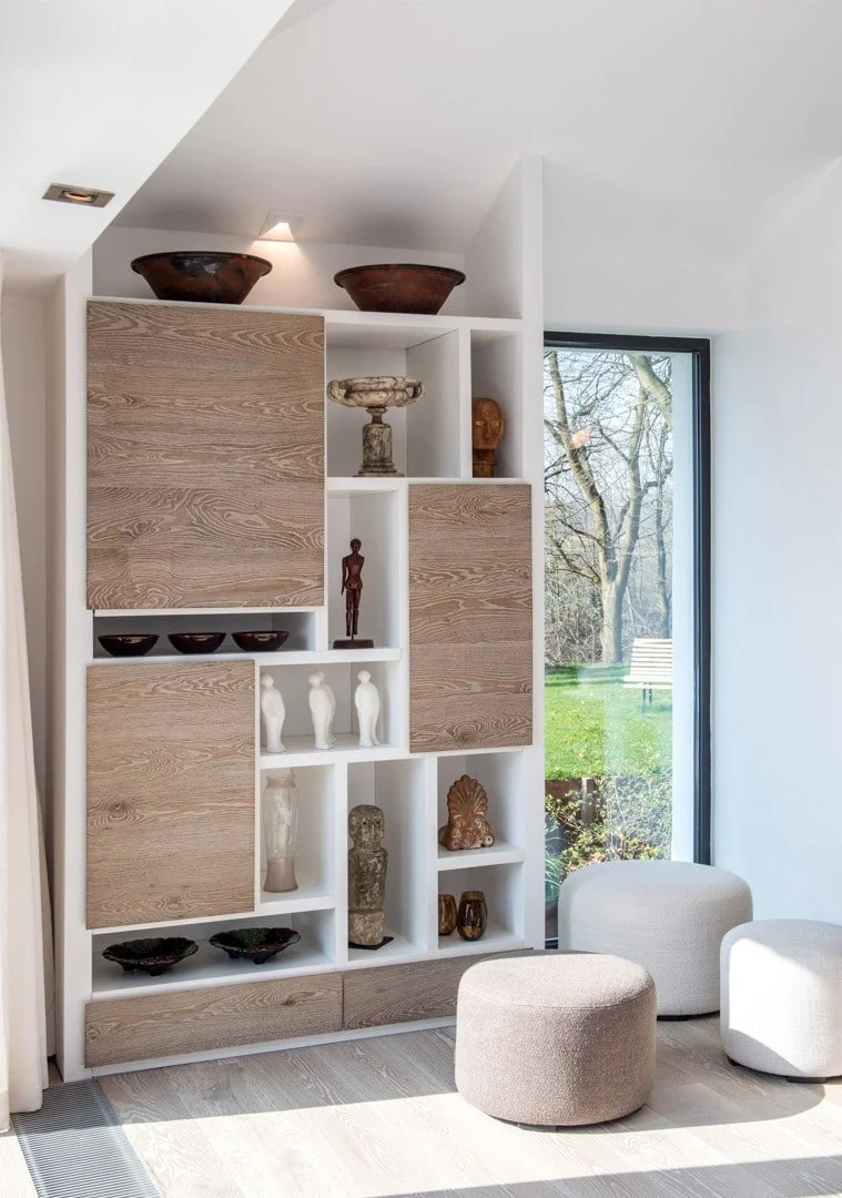 Modern living room corner with a built-in shelving unit displaying decorative vases and sculptures, two beige ottomans, and a large window showing trees and a bench outside.