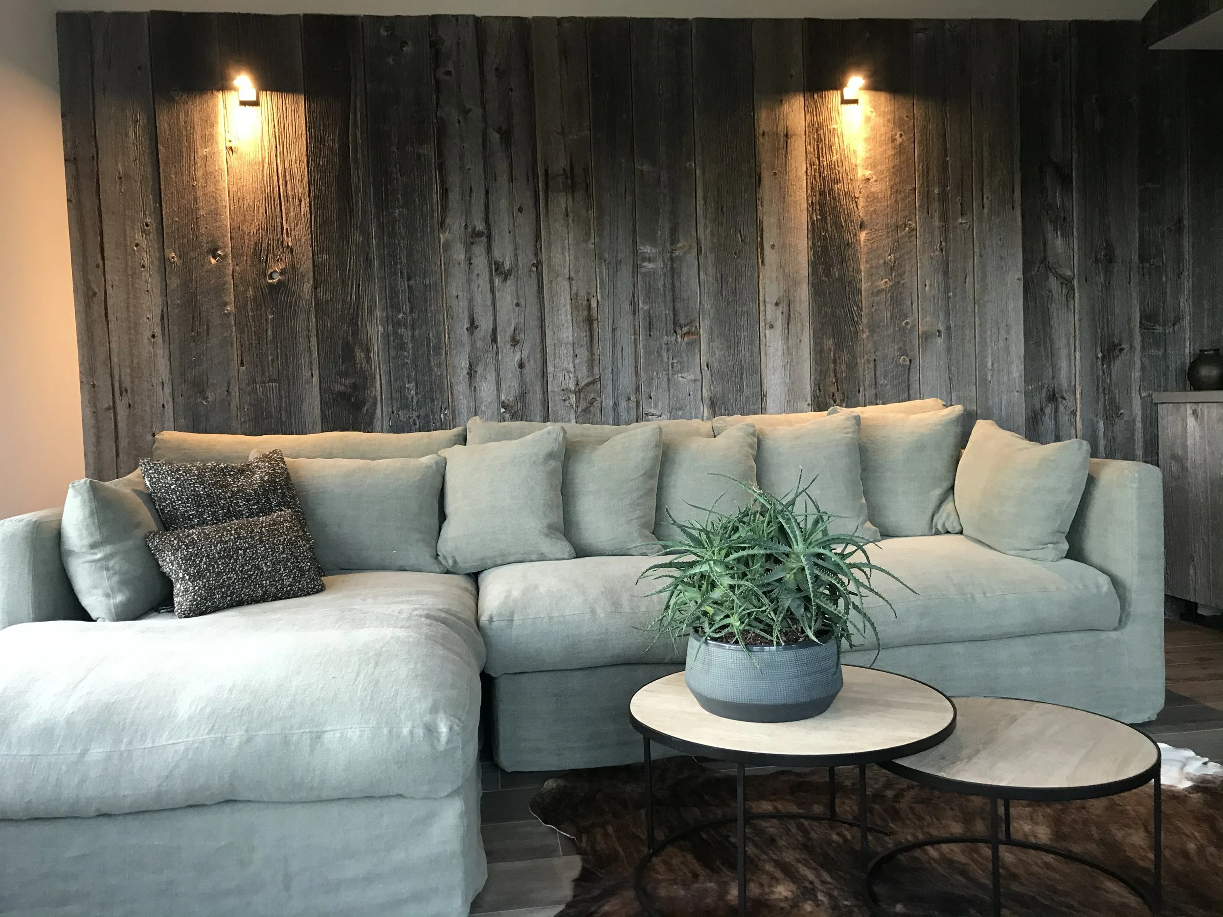 Living room with a large beige sectional sofa adorned with throw pillows, a round coffee table with a two-tone top and a black metal frame, a green potted plant on the coffee table, a wood-paneled accent wall with two wall-mounted lights, and a small side table with a decorative vase.