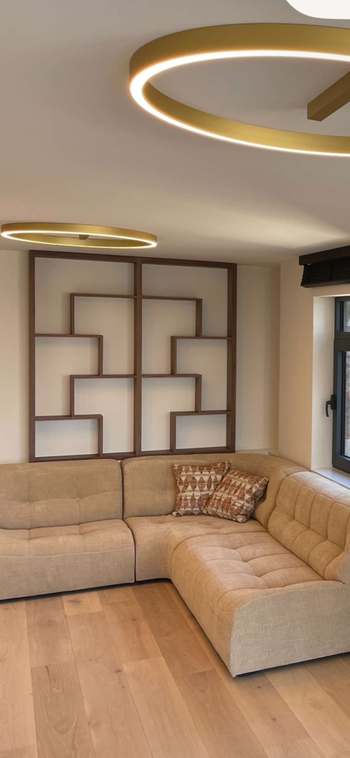 Living room with beige sectional sofa, patterned throw pillows, a modern wooden shelf wall feature, and circular ceiling lights.