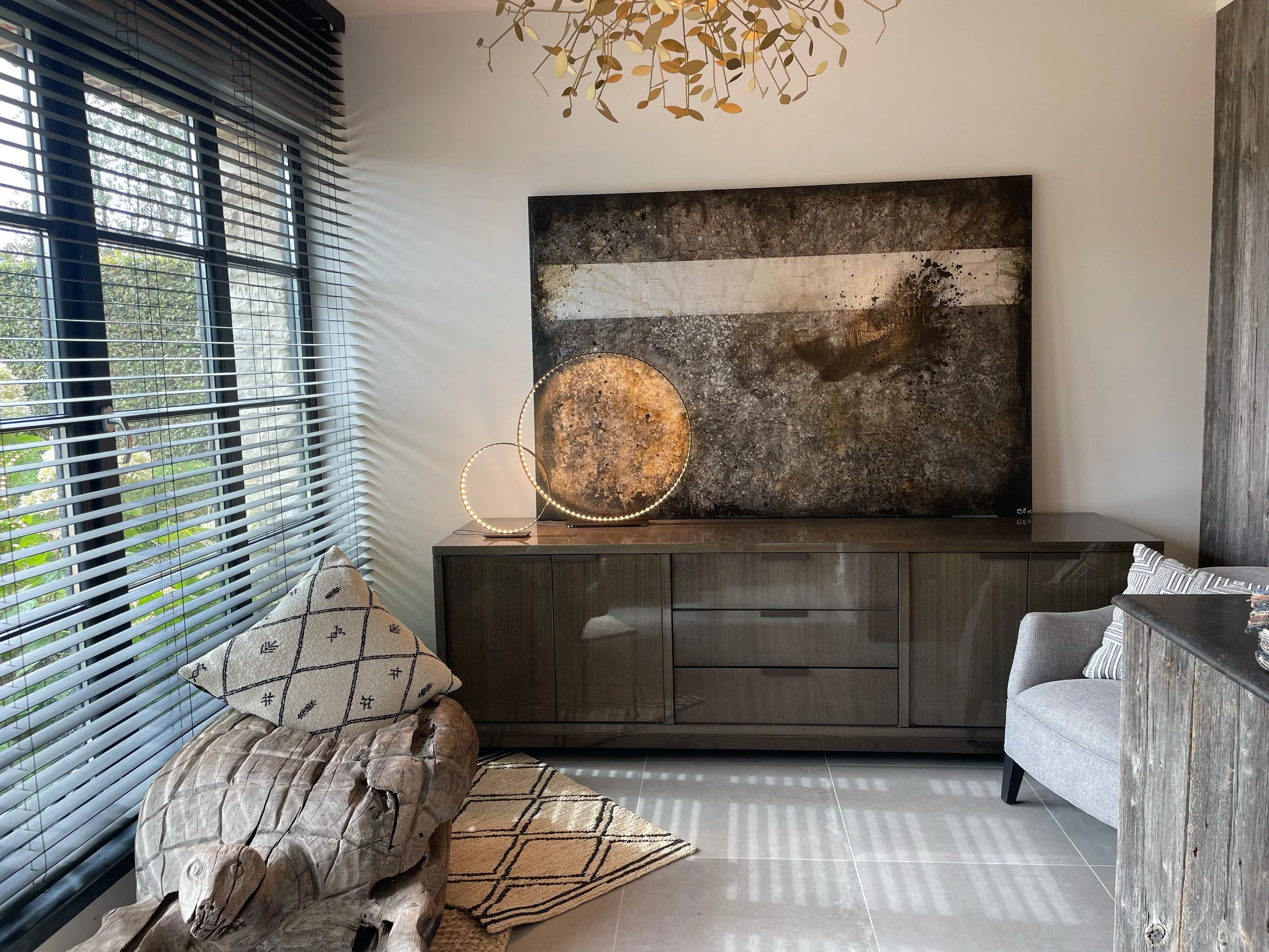 A modern interior room with large windows covered by horizontal blinds, a window seat with a patterned pillow and a piece of furniture made of a large, natural wood log, a dark wood cabinet or sideboard, a geometric rug, a textured wall with an abstract art piece, and a contemporary chandelier with multiple circular LED rings hanging from the ceiling.