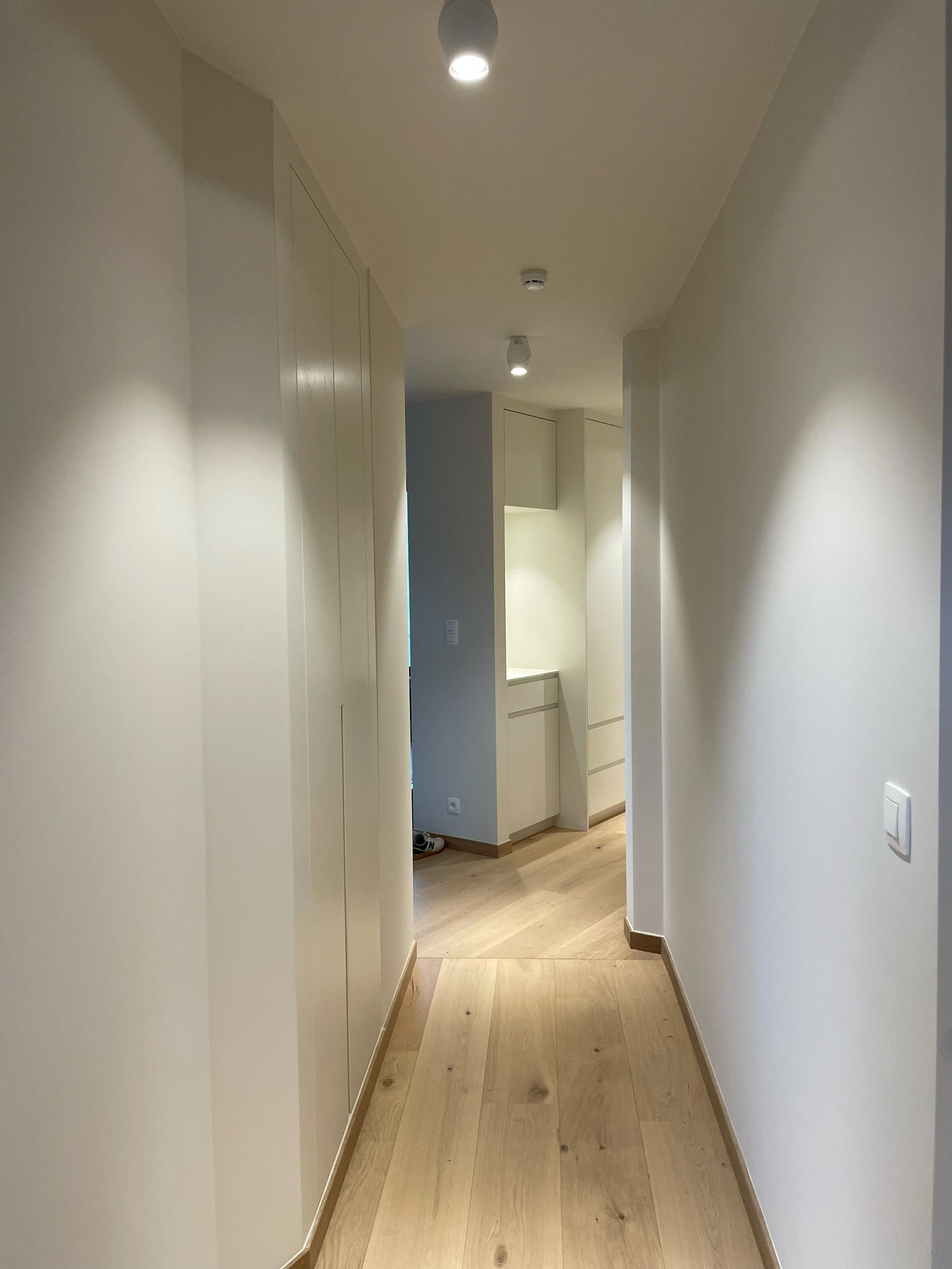 A narrow hallway with light-colored wooden floor, white walls, and a white ceiling with multiple ceiling-mounted lights. There are built-in cabinets along the left side and a small alcove with additional cabinets and drawers at the end of the hallway.