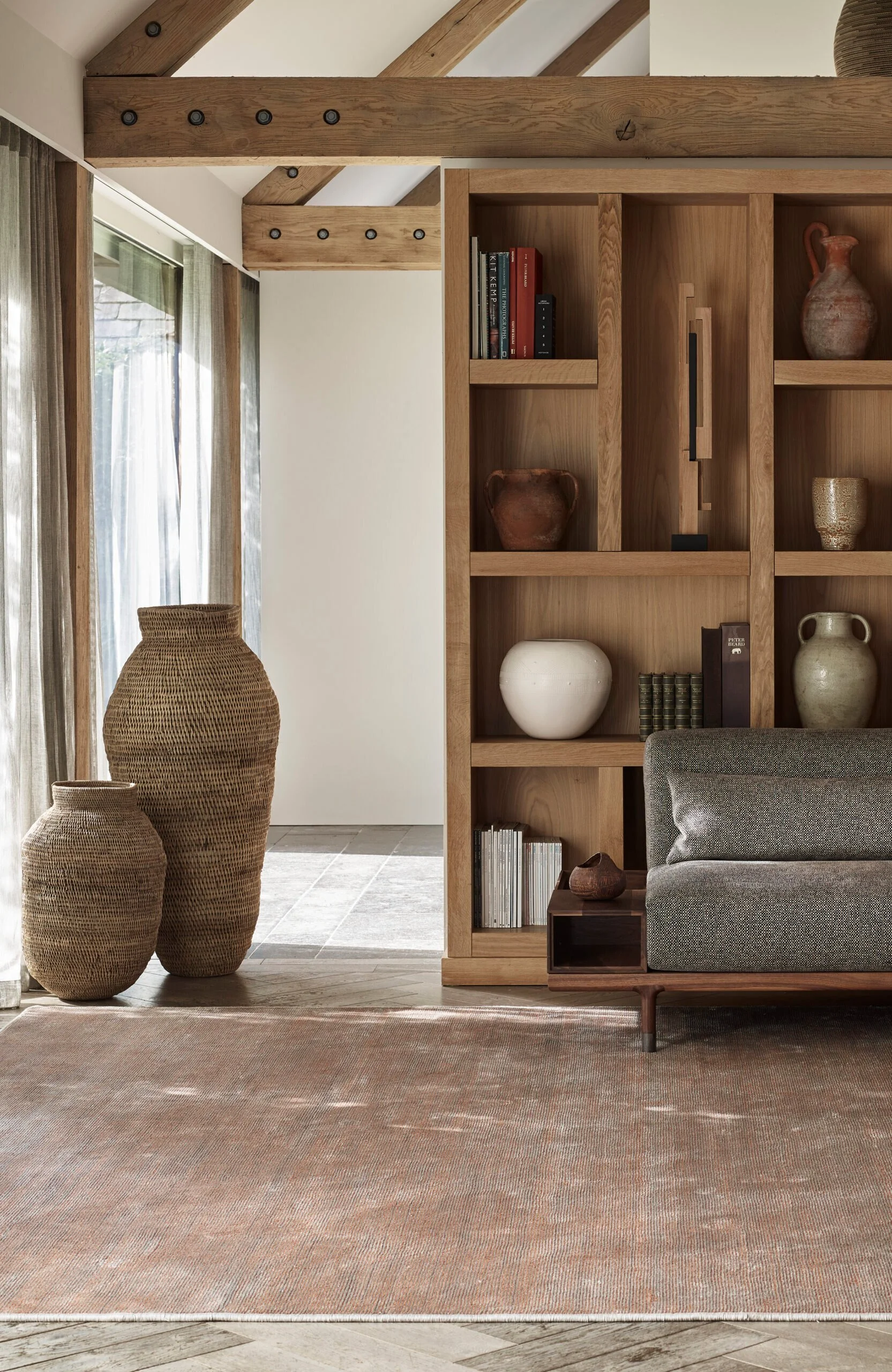 Interior of a modern living room with a wooden bookshelf filled with ceramics, books, and decor, a large woven basket, a grey sofa, and a beige area rug on a wood and tile floor.