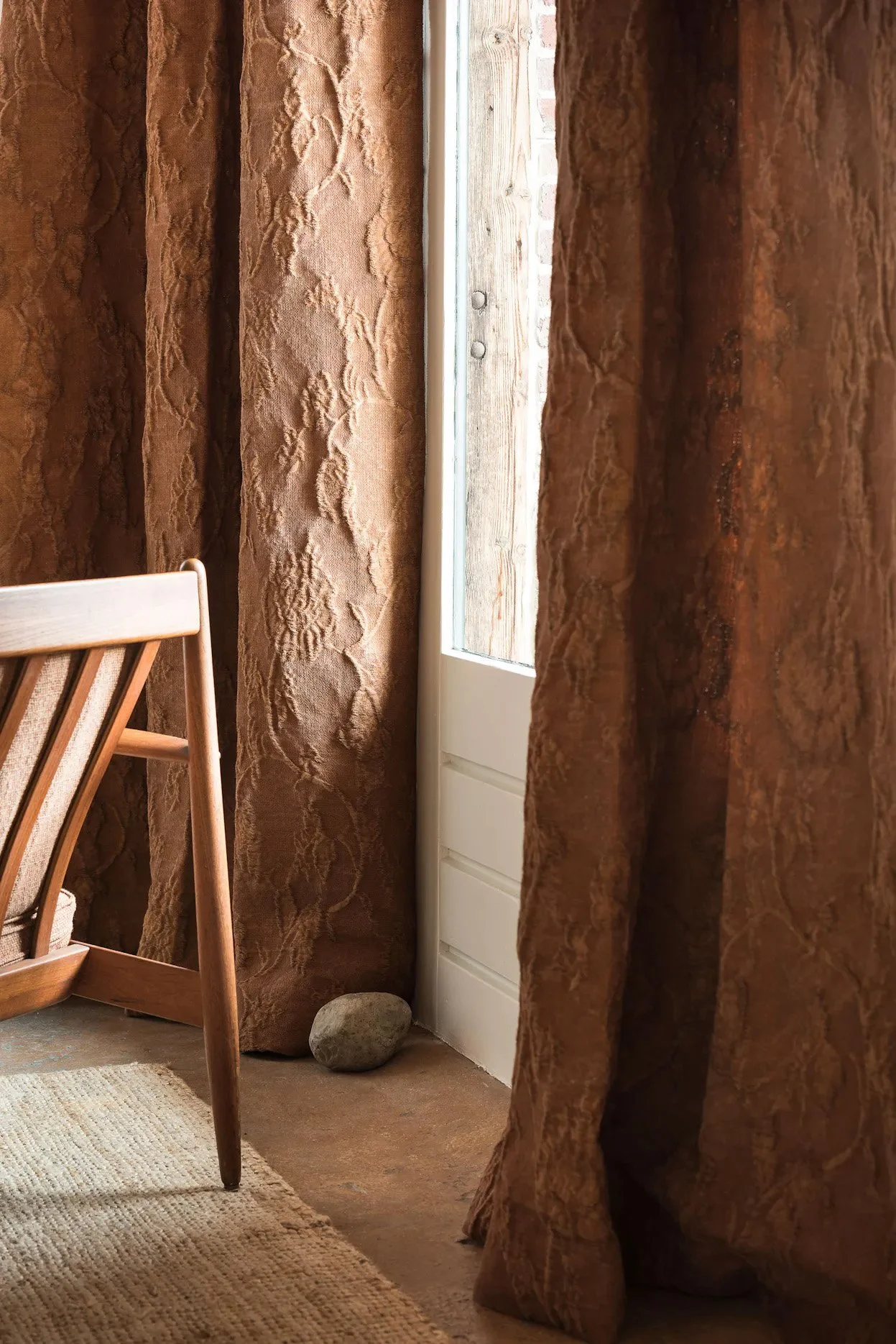 A corner of a room with brown patterned curtains, a wooden chair, a small stone on the floor, and a window with a view of a brick wall outside.