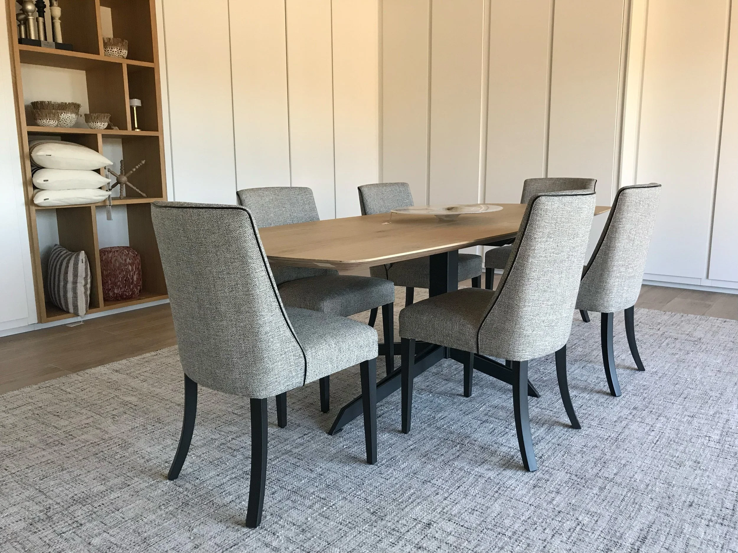 Interior of a dining area with a wooden table surrounded by six upholstered chairs. In the background, there is a wall with vertical panels and a wooden shelving unit with bowls, pillows, and decorative items. A circular tray is on the table, and a rug covers the floor beneath the table.