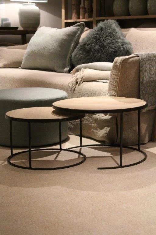 Two nested oval coffee tables with black metal frames and light wood tops in front of a beige sofa with pillows and a grey furry throw blanket.