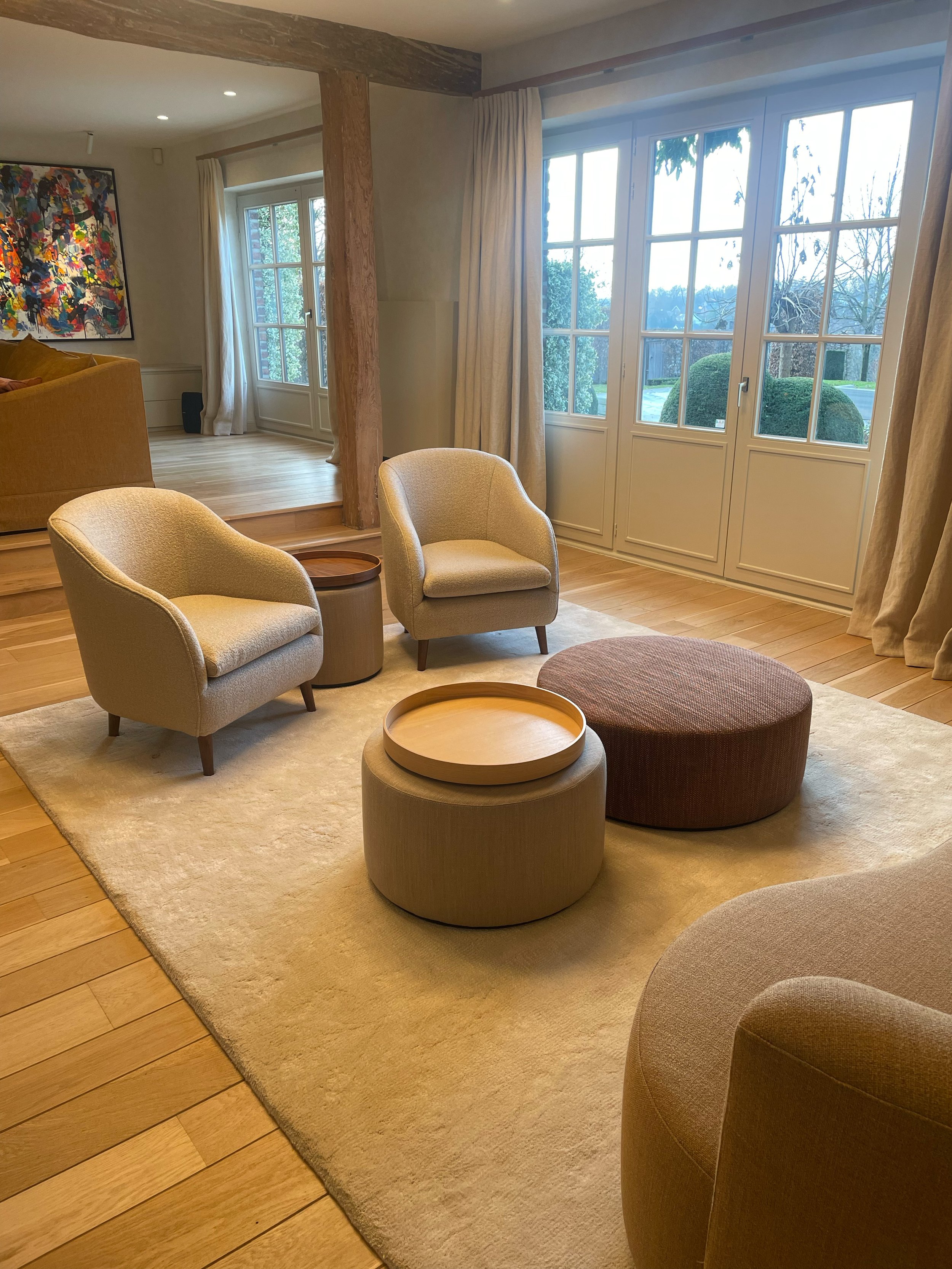 Living room with large windows, beige chairs, a round ottoman, a beige rug, and artwork on the wall.