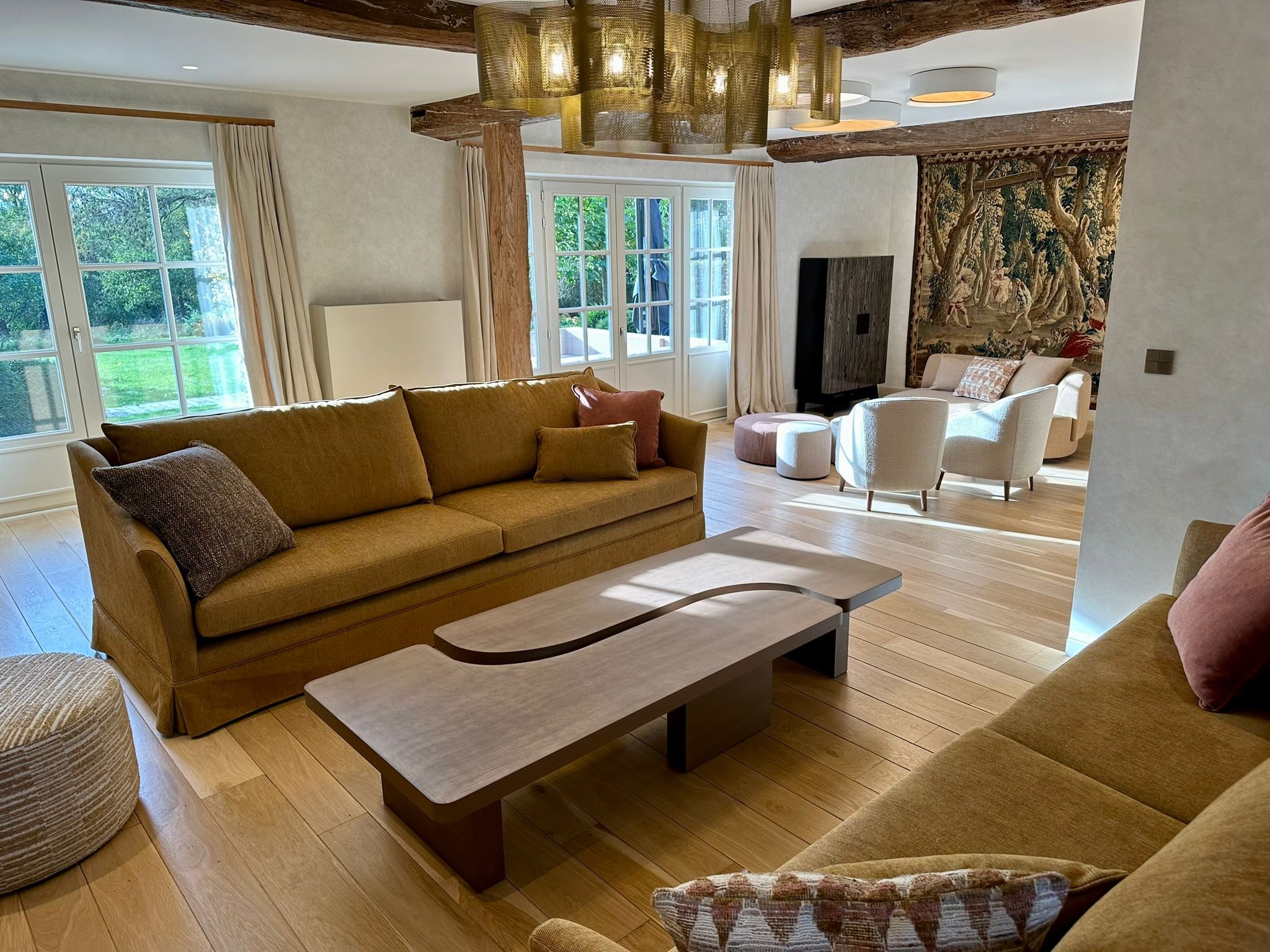 A cozy living room with a yellow sofa, a wooden coffee table, and a large window with cream curtains. There is a wall tapestry, a TV, and a white armchair with a patterned pillow. The ceiling has exposed wooden beams and modern light fixtures, and the floor is made of light wood.