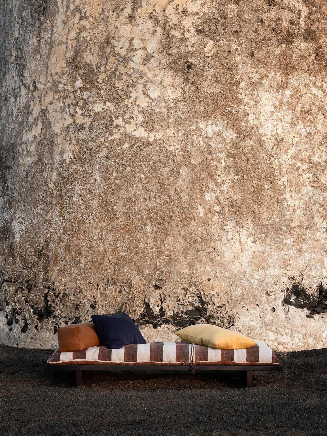A long striped bed with several pillows against a weathered, textured wall. The bed is on a gravel ground.