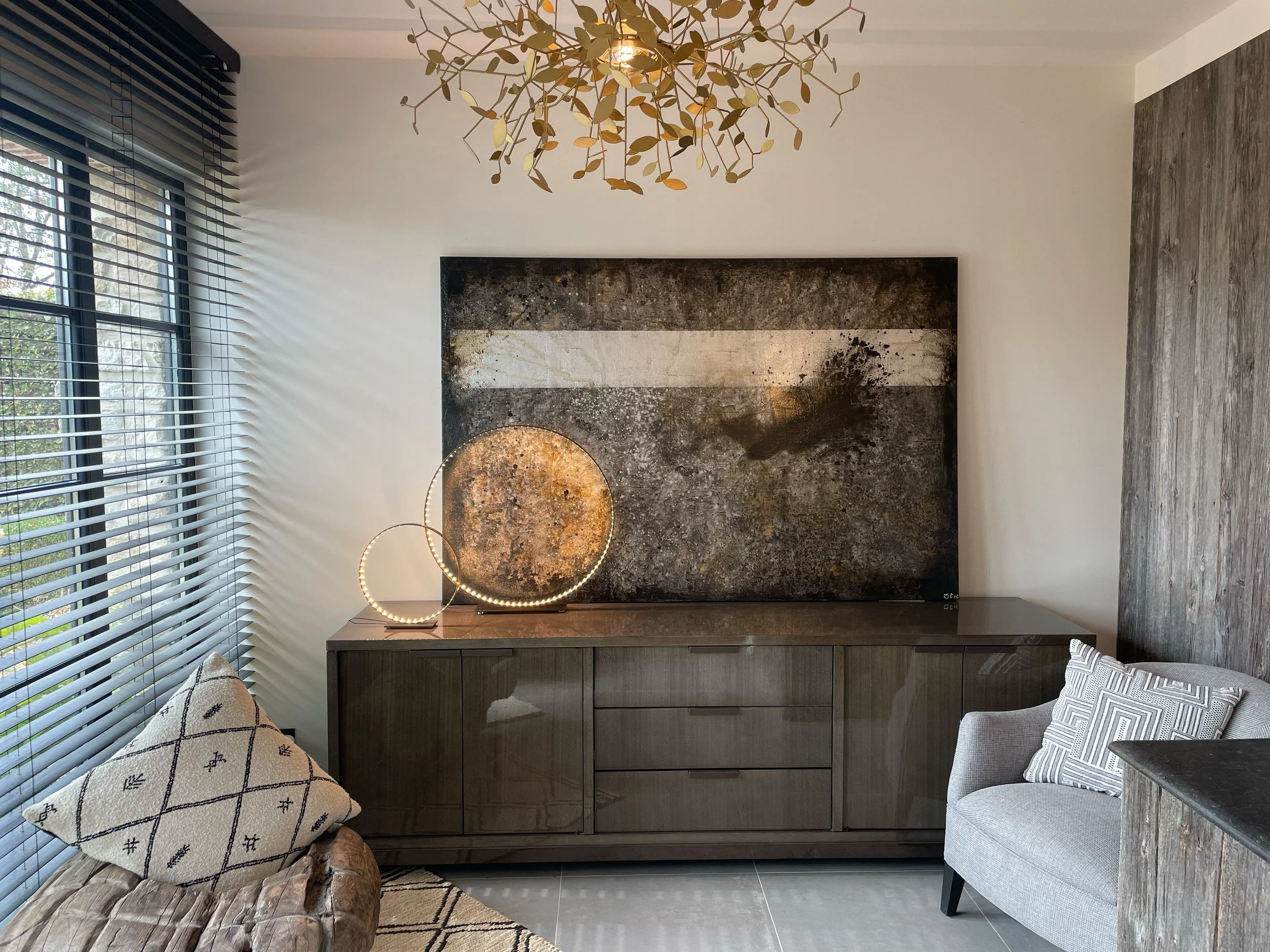 Modern living room with large window, black horizontal blinds, marble fireplace with gray cabinets below, two circular LED pendant lights, gray upholstered armchair, decorative pillows, textured wooden side table with blanket, hardwood floor, and abstract art on the wall.