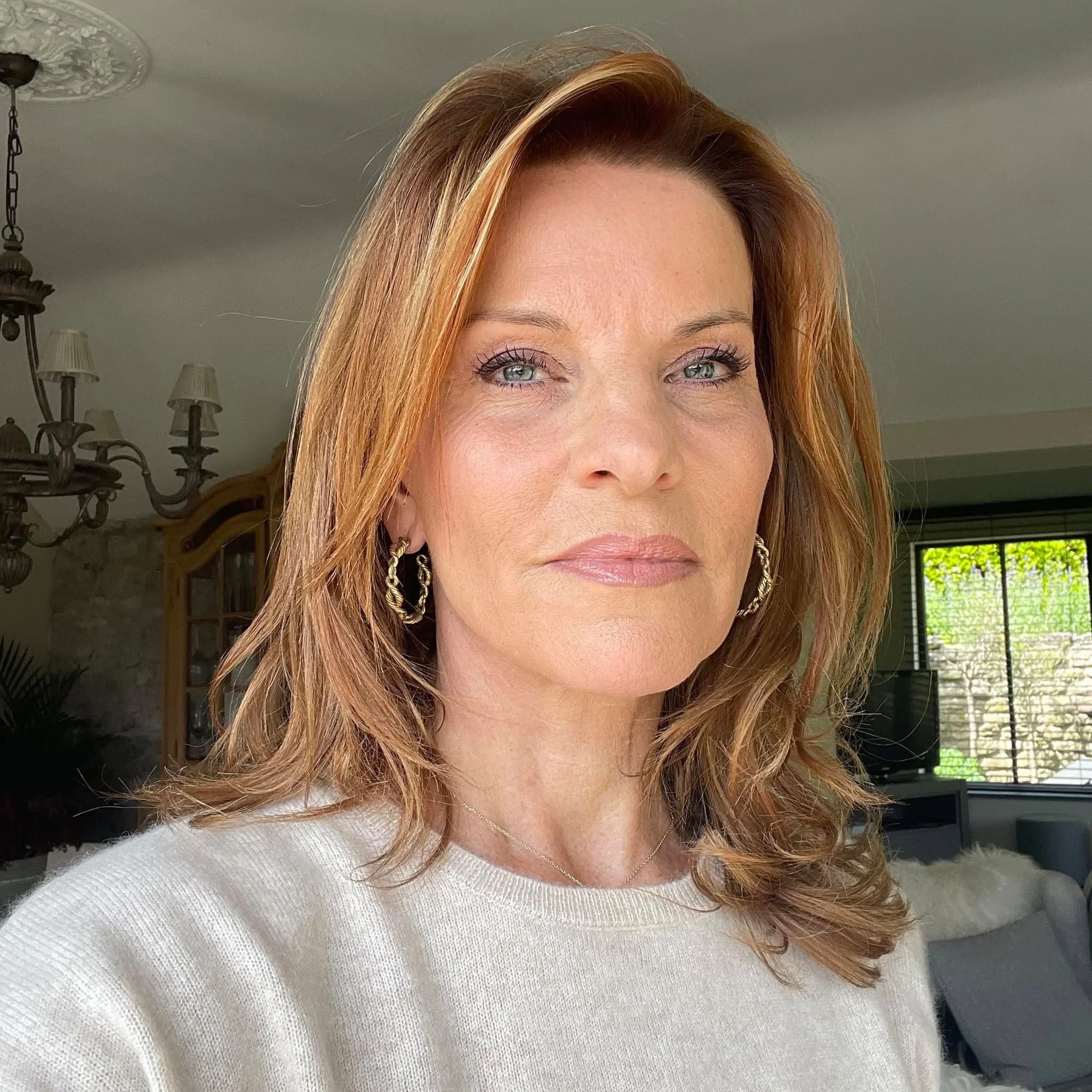 Close-up of a woman with blue eyes and reddish-brown hair wearing earrings and a cream-colored sweater, indoors with a chandelier and settlement items in the background.
