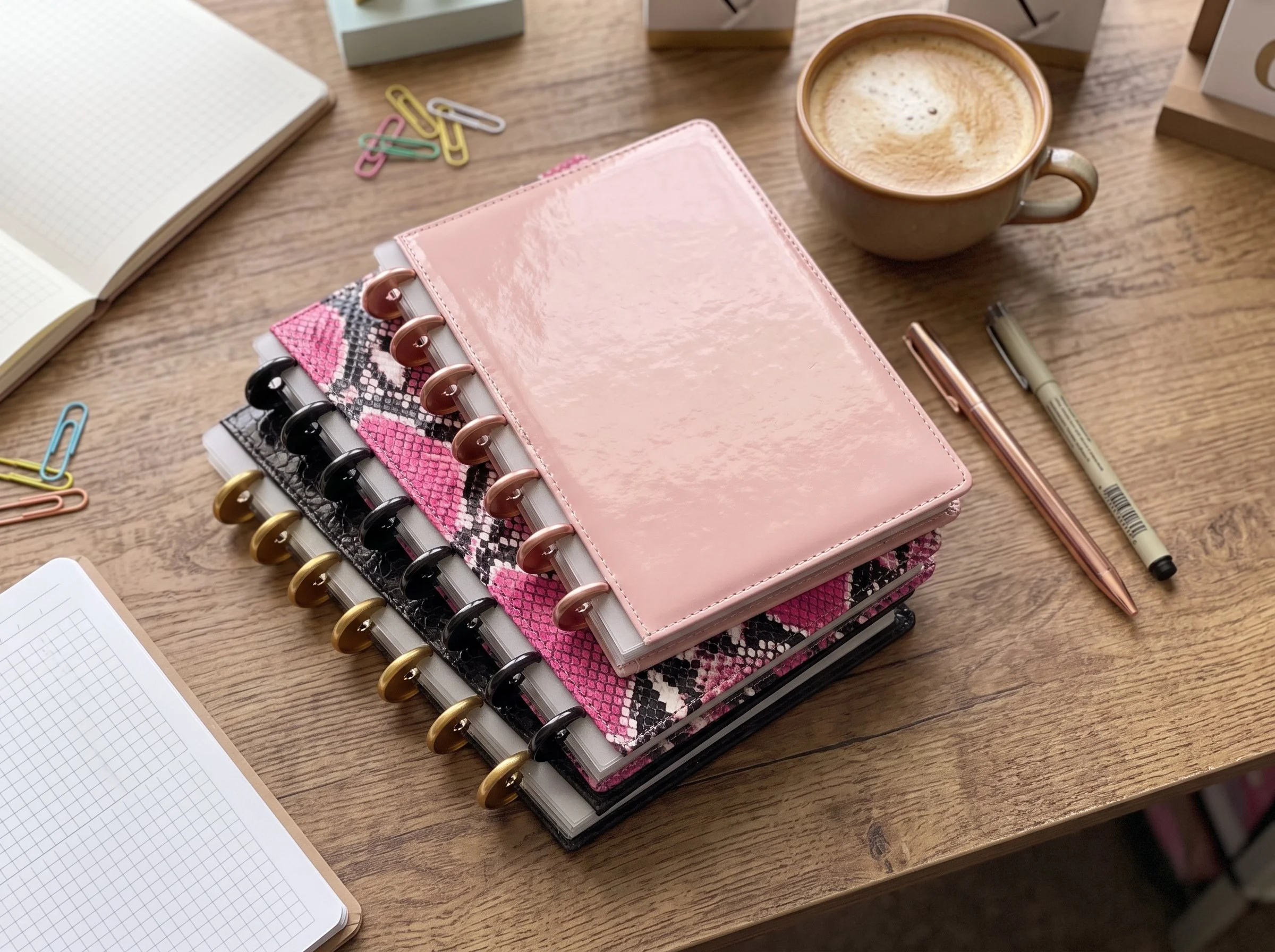 A wooden desk with a stack of notebooks and planners, a cup of coffee, a pen, paper clips, and an open notebook with graph paper.