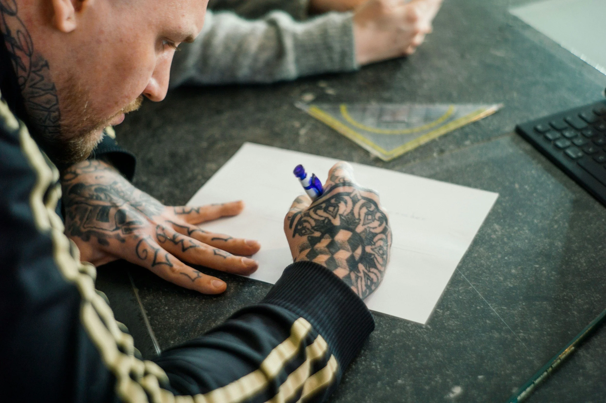A person with tattoos on arms and neck writing on a sheet of white paper with a blue marker, sitting at a desk with a computer keyboard and rulers.