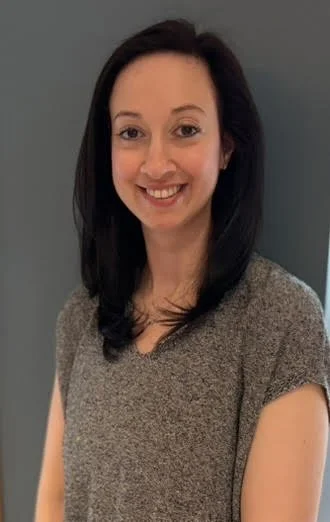 A woman with long dark hair smiling, wearing a gray short-sleeve top, standing in front of a gray wall.