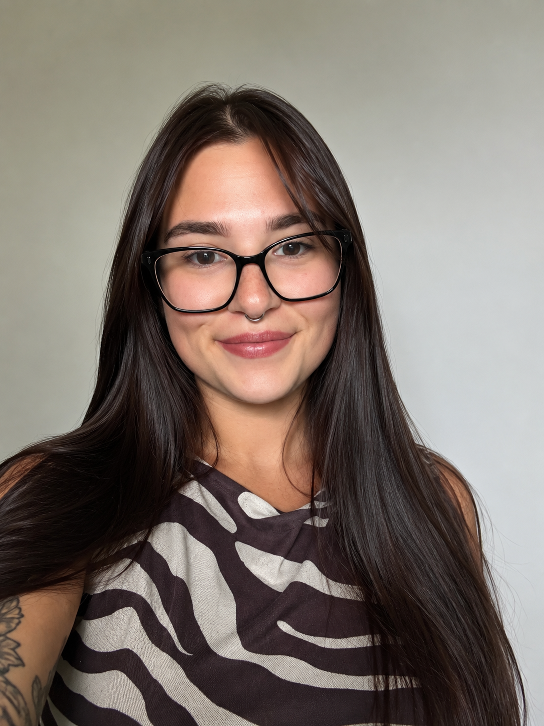 Portrait of a young woman with long dark hair, wearing black glasses and a zebra print sleeveless top, smiling against a plain background.