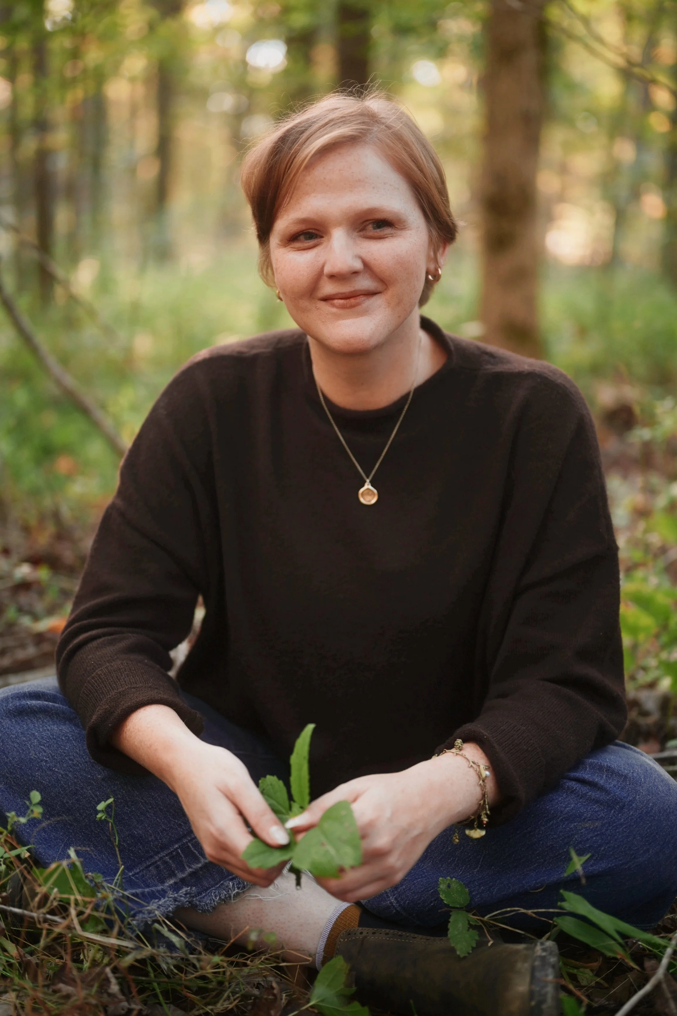 A young woman with short red hair, freckles, and earrings, sitting outdoors in a forest, planting a small green sapling while smiling at the camera.