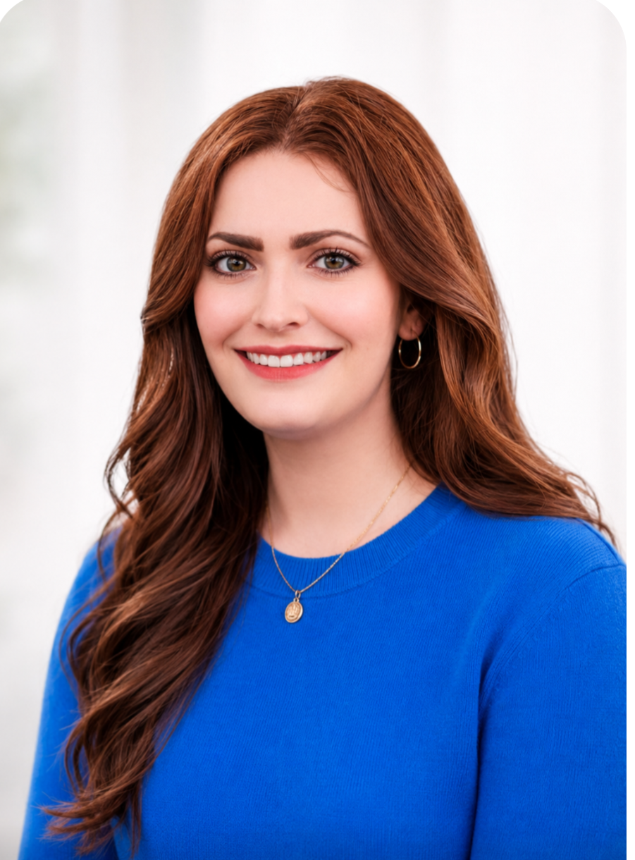 A woman with long, wavy red hair and green eyes, smiling, wearing a blue top, gold hoop earrings, and a gold necklace with a small pendant, standing against a plain light background.