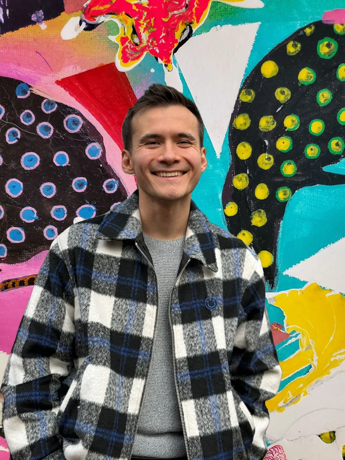 A smiling young man standing in front of a colorful abstract mural painting with black butterflies, dots, and vibrant colors.